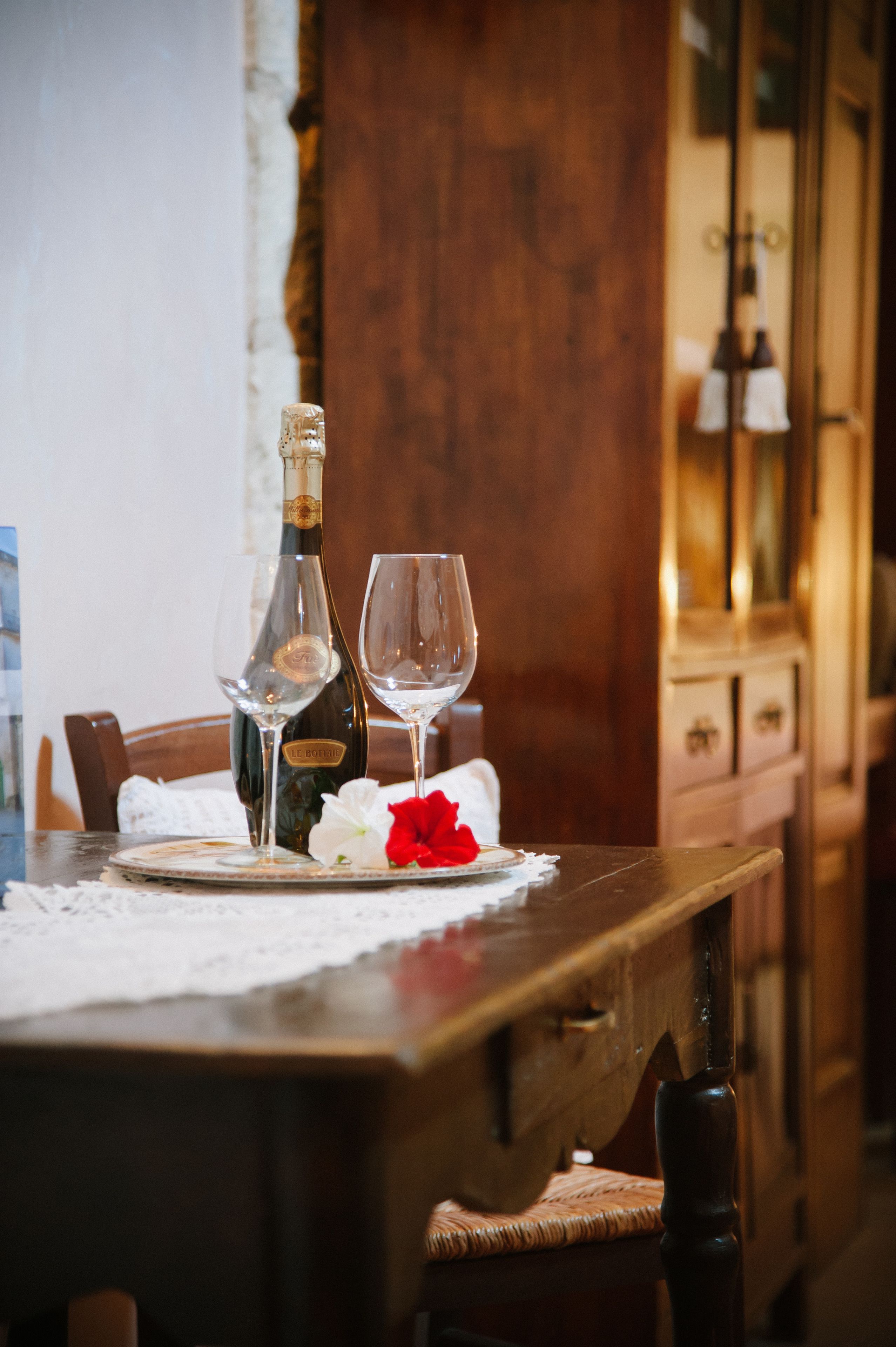 A dining table with a bottle and two wine glasses, a red rose, and a lace tablecloth.