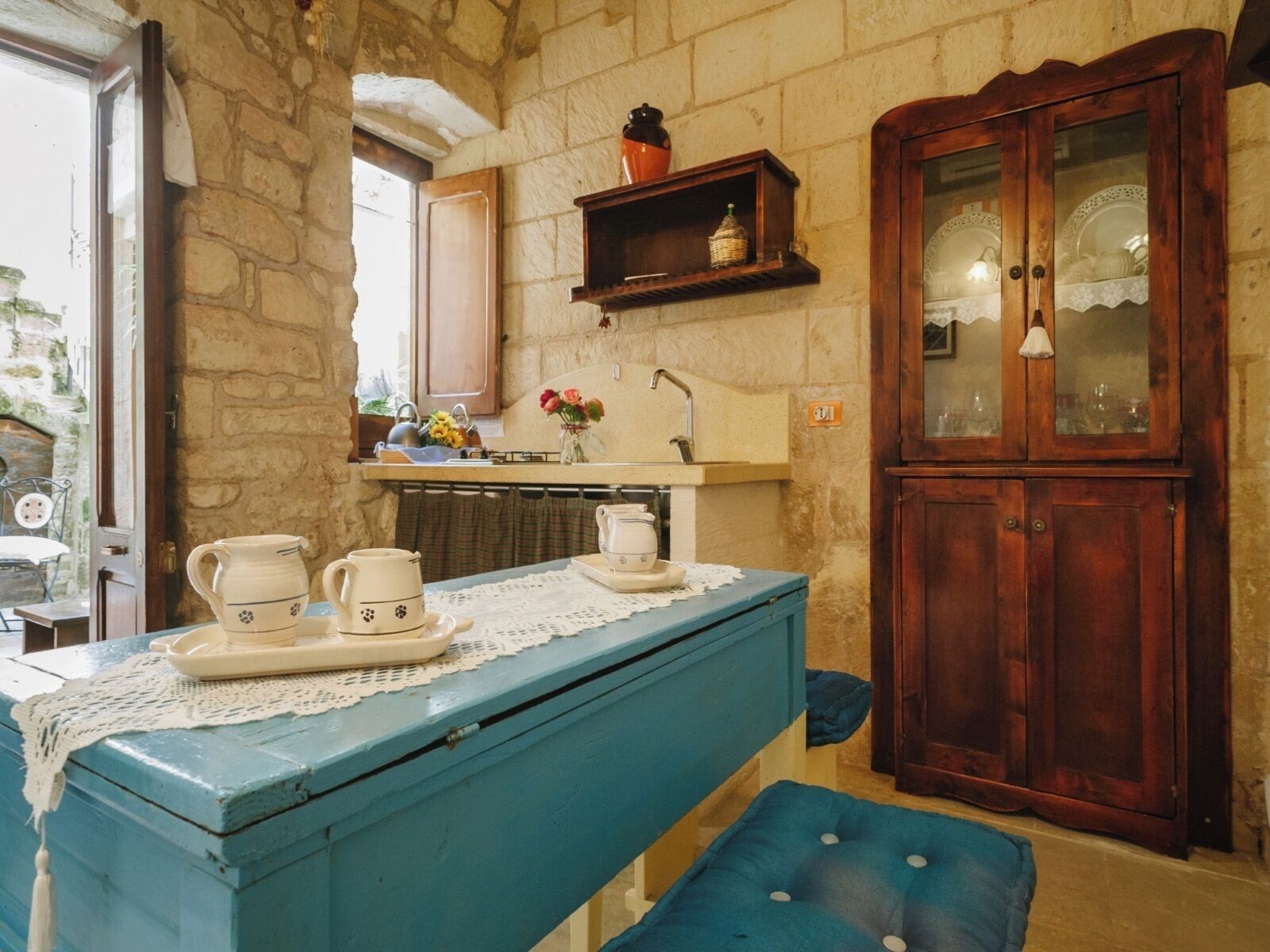 A rustic kitchen with a stone wall, a wooden cabinet with glass doors, a blue table with tea sets, and a window overlooking a courtyard.