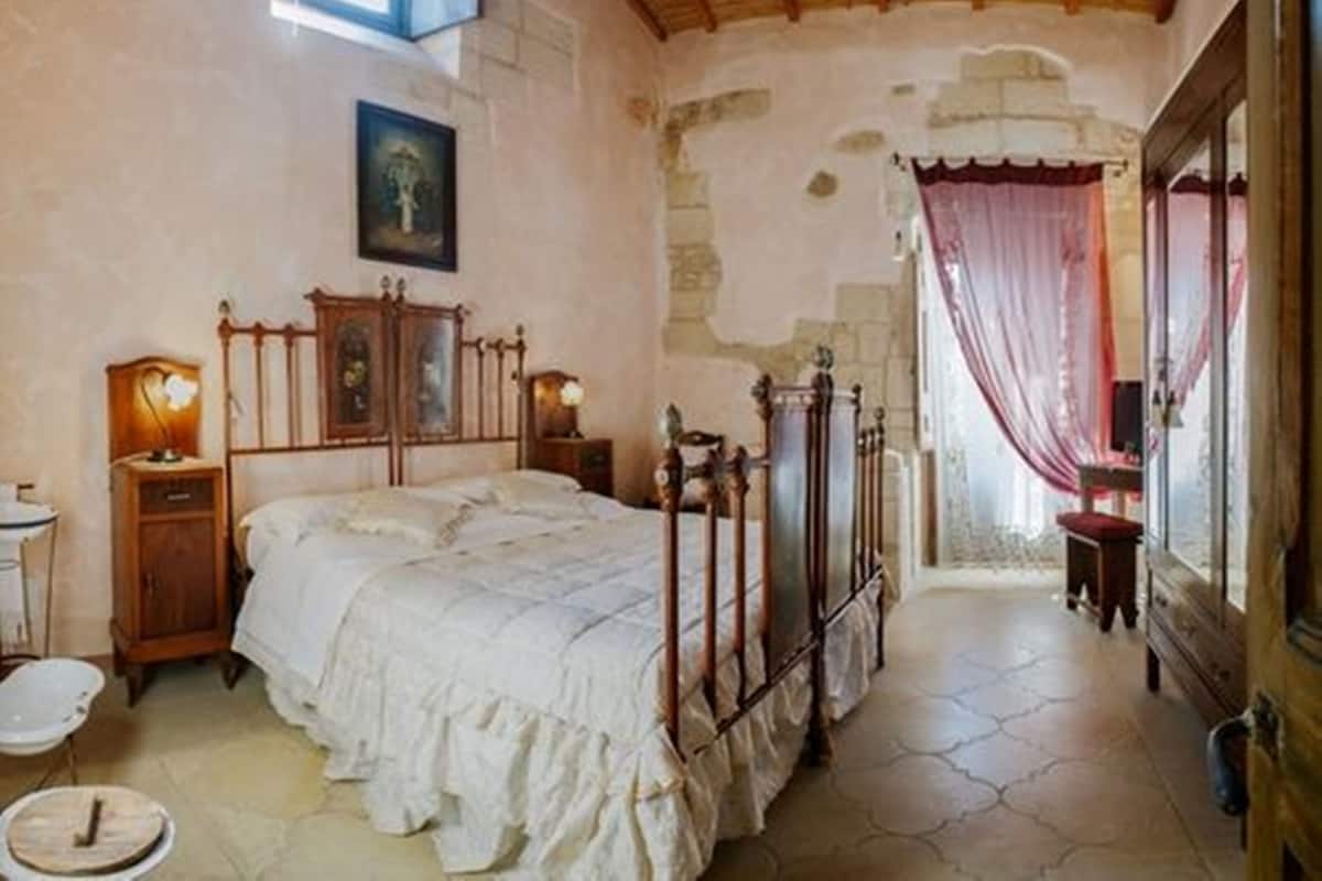 Ein Schlafzimmer mit einem hölzernen Bett, einem Nachttisch, einem Fenster mit Vorhängen und einem Steinboden.