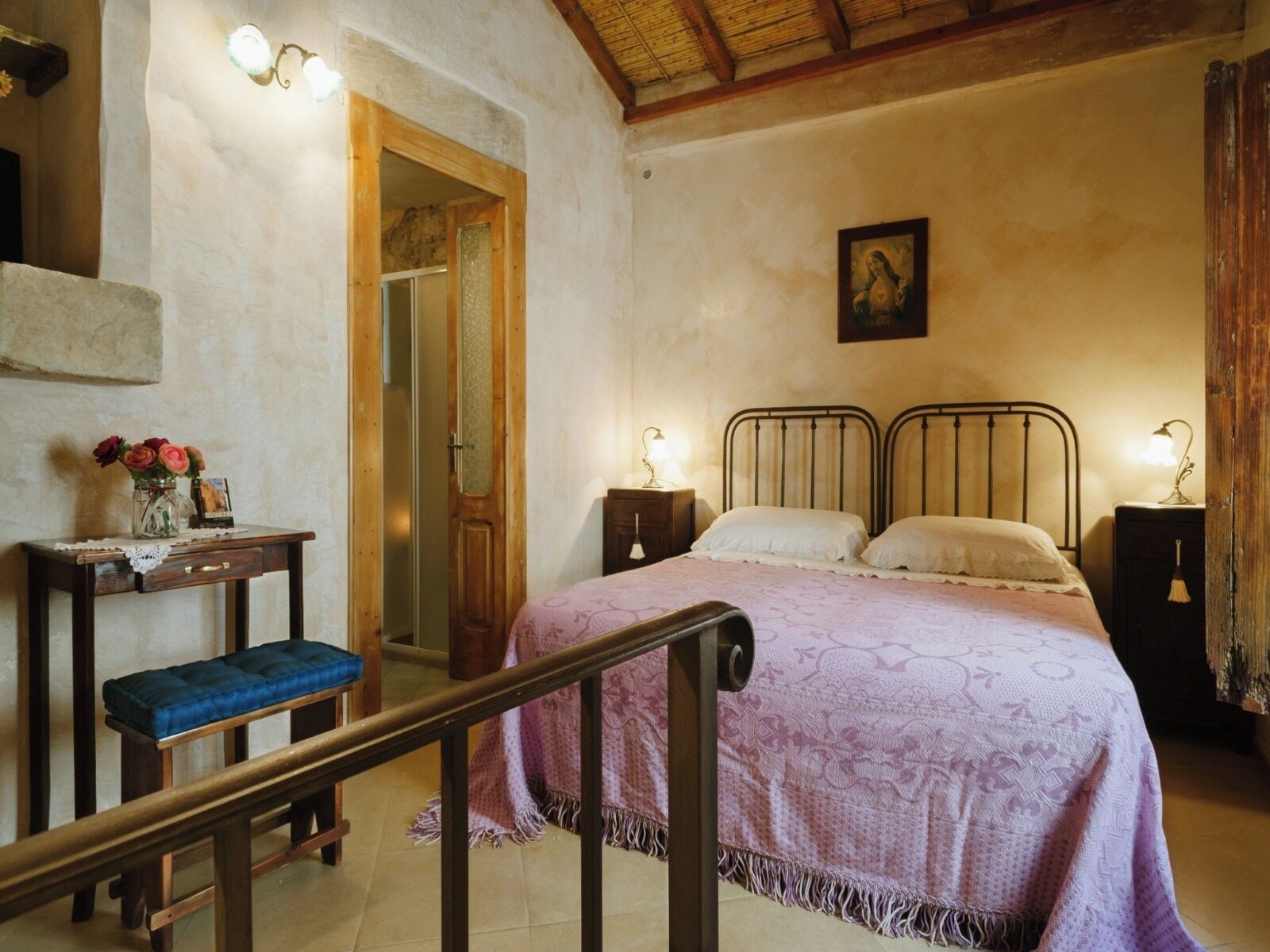 A bedroom with a bed, bedside tables, a bench, and a door leading to another room.