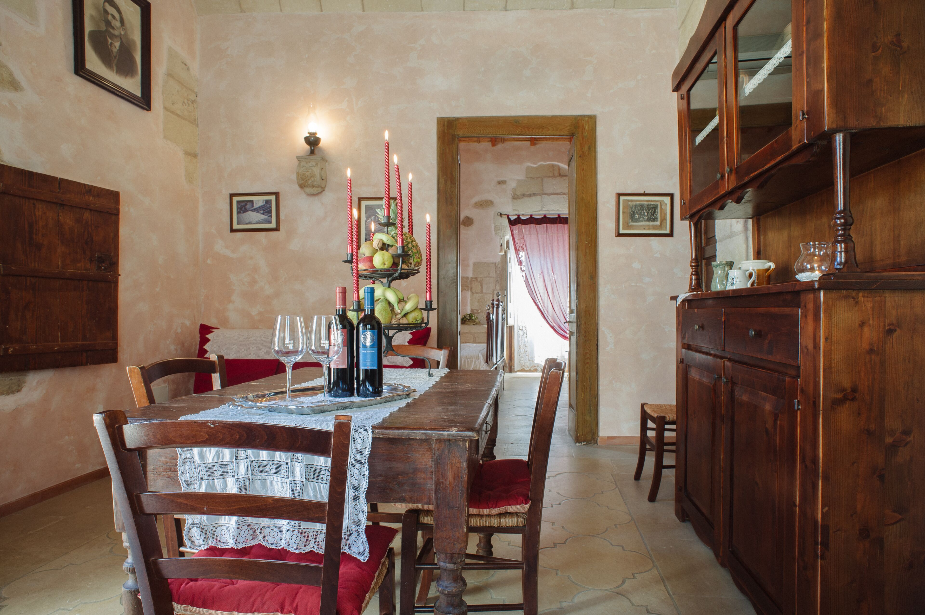 Zona pranzo con tavolo in legno apparecchiato per quattro, circondato da sedie con cuscini rossi. Ci sono candele sul tavolo, e sullo sfondo si nota un vassoio di frutta.