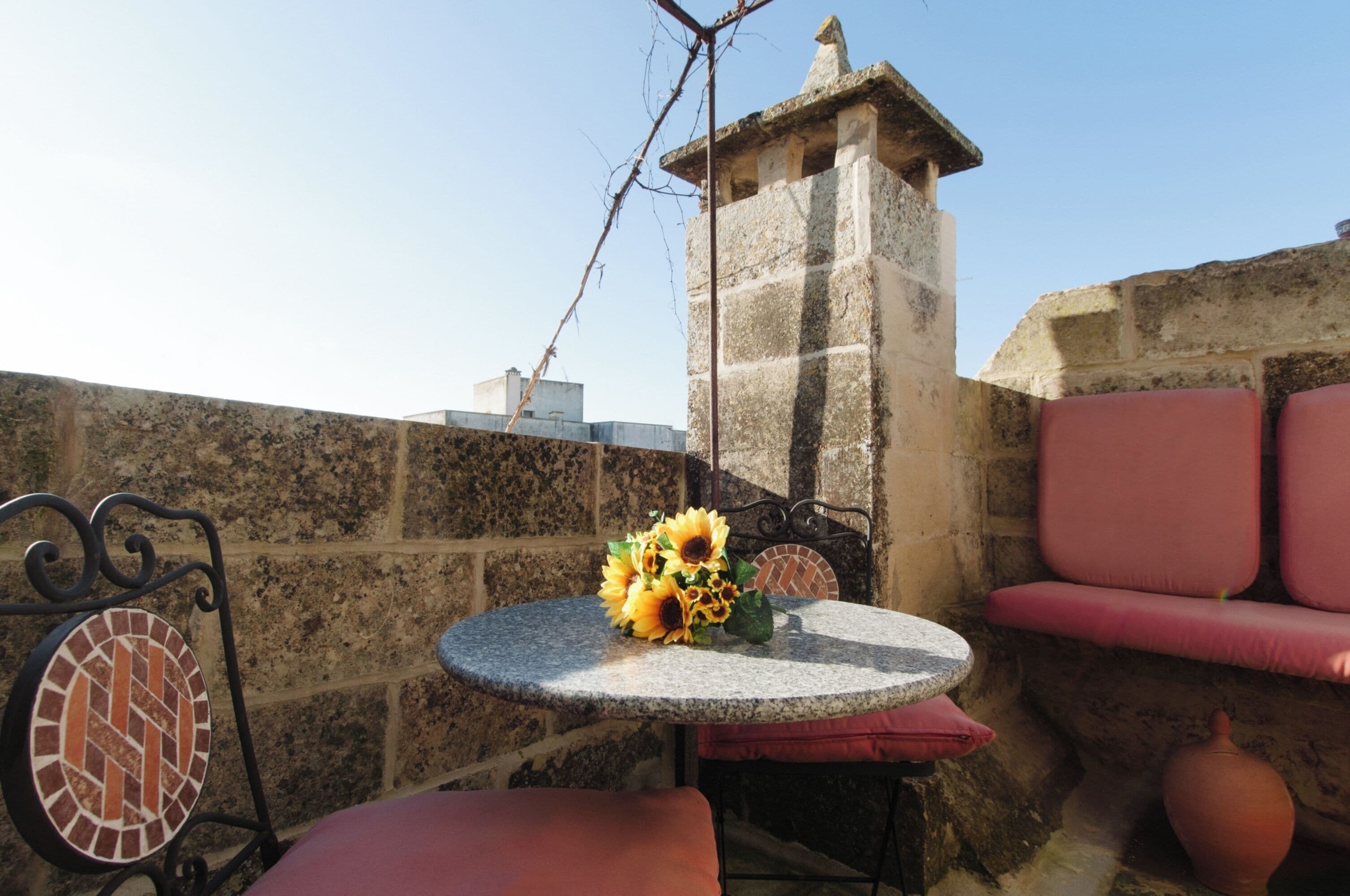 Un muro de piedra, una mesa redonda con un jarrón de girasoles, una silla de metal y un banco acolchado.
