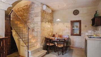A stone-walled kitchen with a dining table, chairs, a wooden staircase, and a small window.
