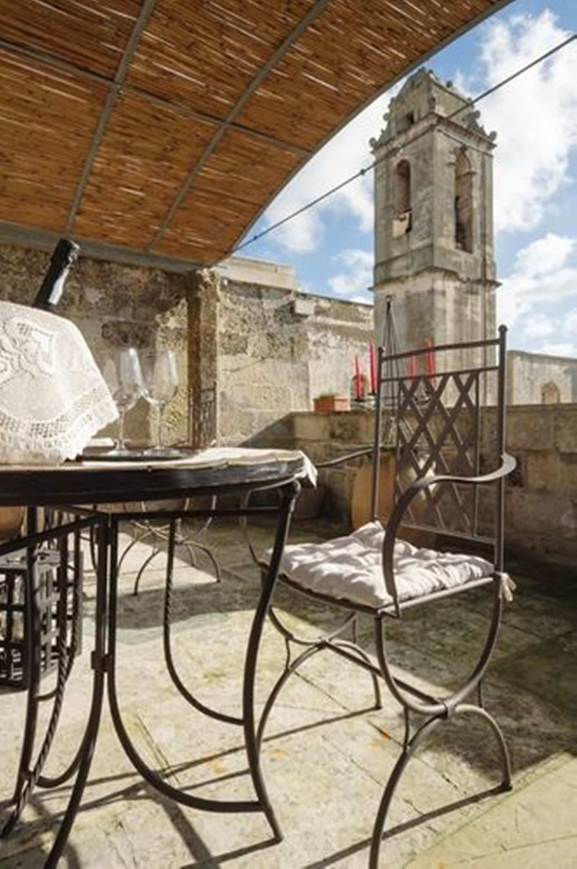 Patio esterno con tavolo e sedia, soffitto in legno e parete in pietra, sullo sfondo un campanile.