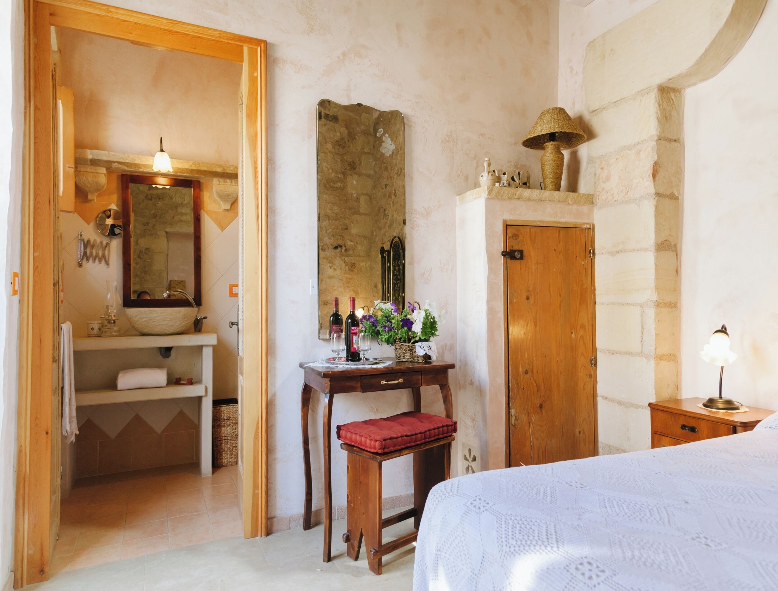 Un dormitorio con una cama de madera, una mesita de noche con lámpara, un escritorio de madera con espejo, y un baño con lavabo y espejo.