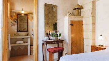 Un dormitorio con una cama de madera, una mesita de noche con lámpara, un escritorio de madera con espejo, y un baño con lavabo y espejo.