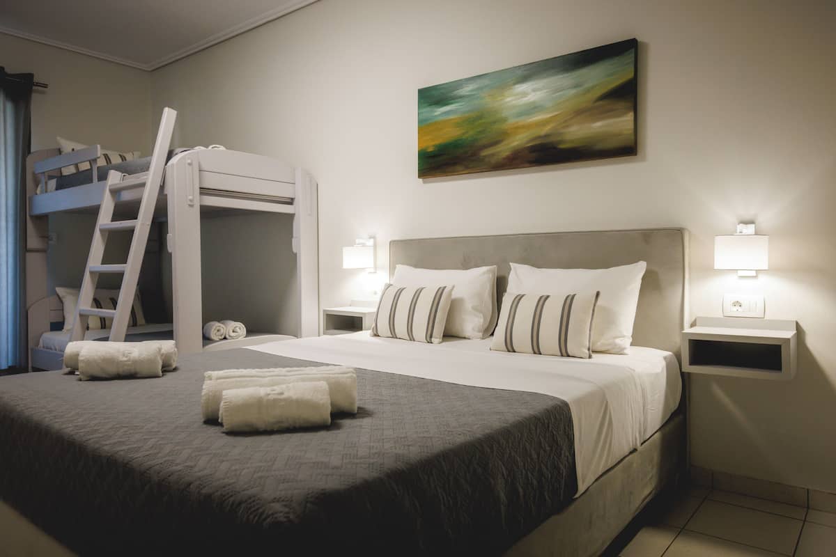 A hotel room with a bunk bed, a bedside table with a lamp, a painting on the wall, and a neatly made bed with towels.