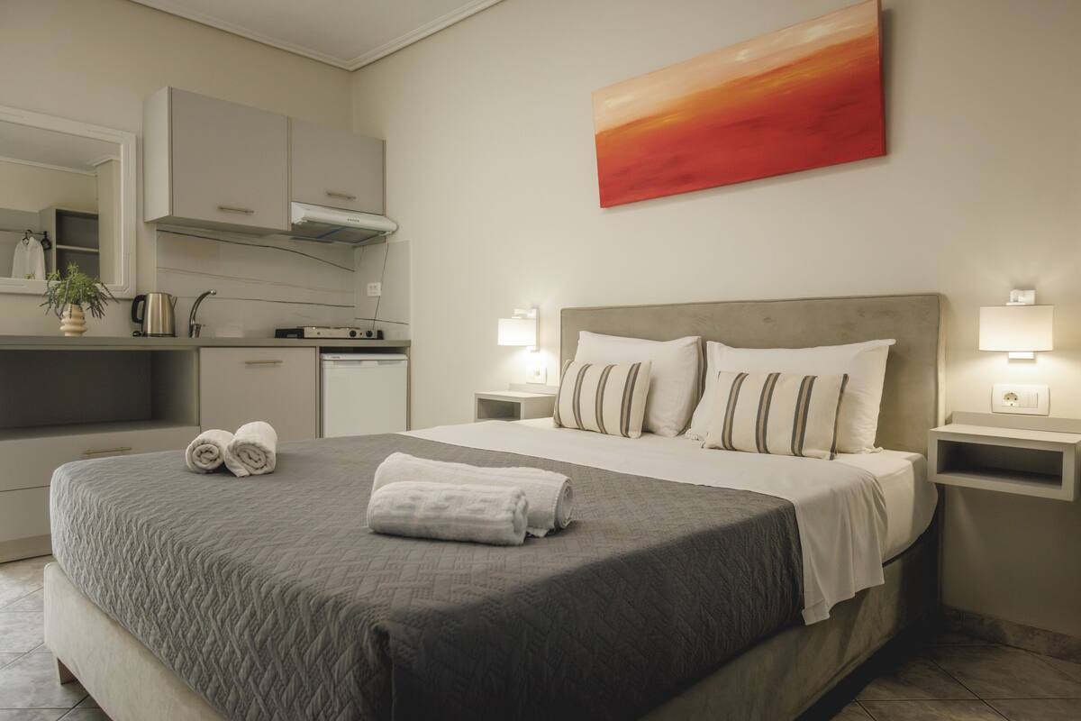 A hotel room with a bed, bedside tables, a nightstand, a mirror, a painting, and a kitchenette.