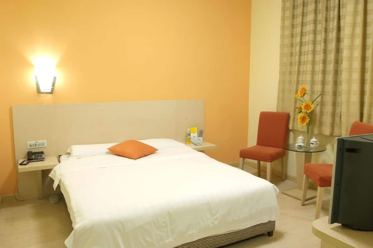 Quarto de hotel com uma cama, uma escrivaninha com telefone, uma cadeira, uma mesinha com bule e chá, e uma televisão.