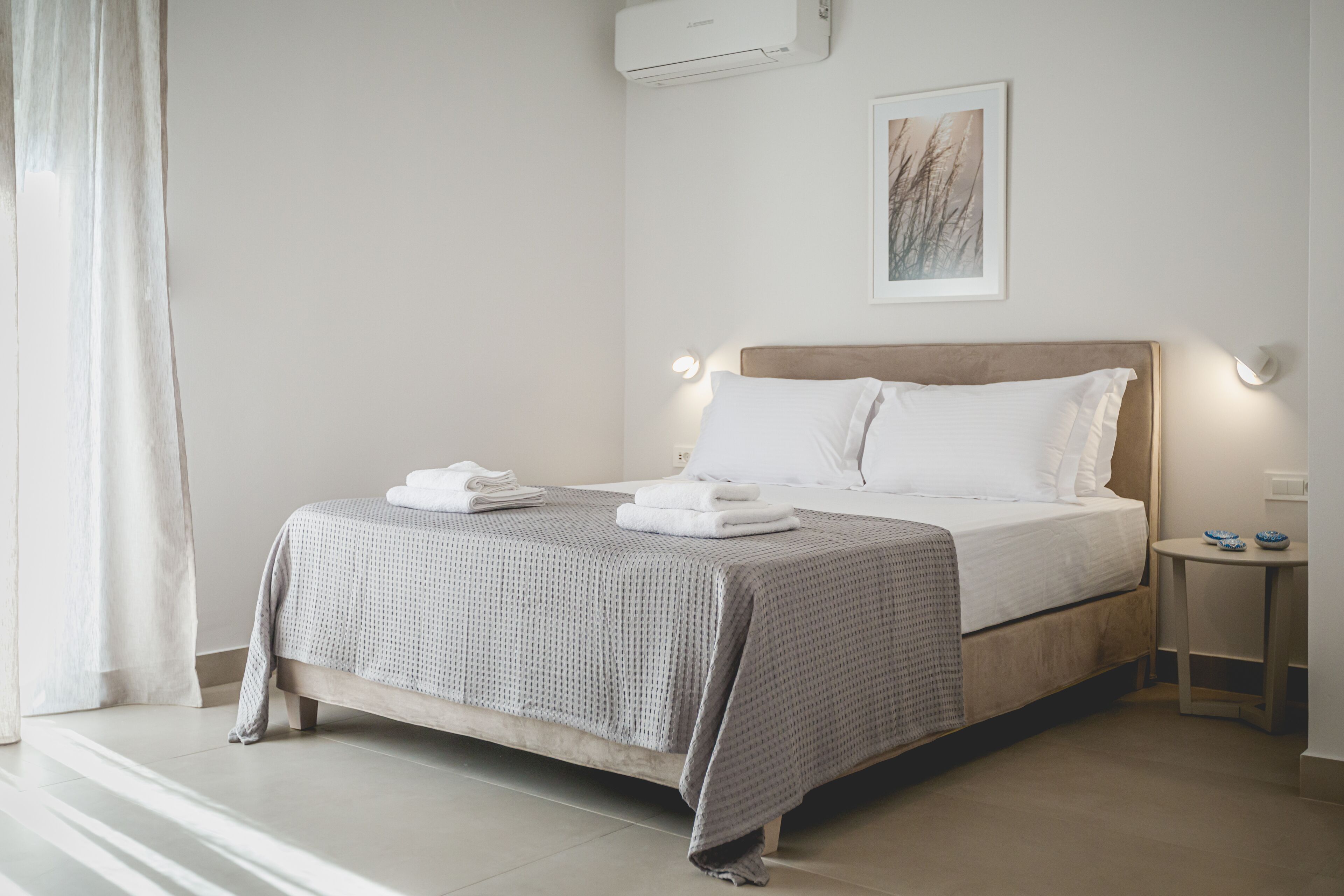 Una cama bien hecha con sábanas blancas y una manta gris estampada, una mesita de noche con un cuenco azul, y una obra de arte enmarcada en la pared.