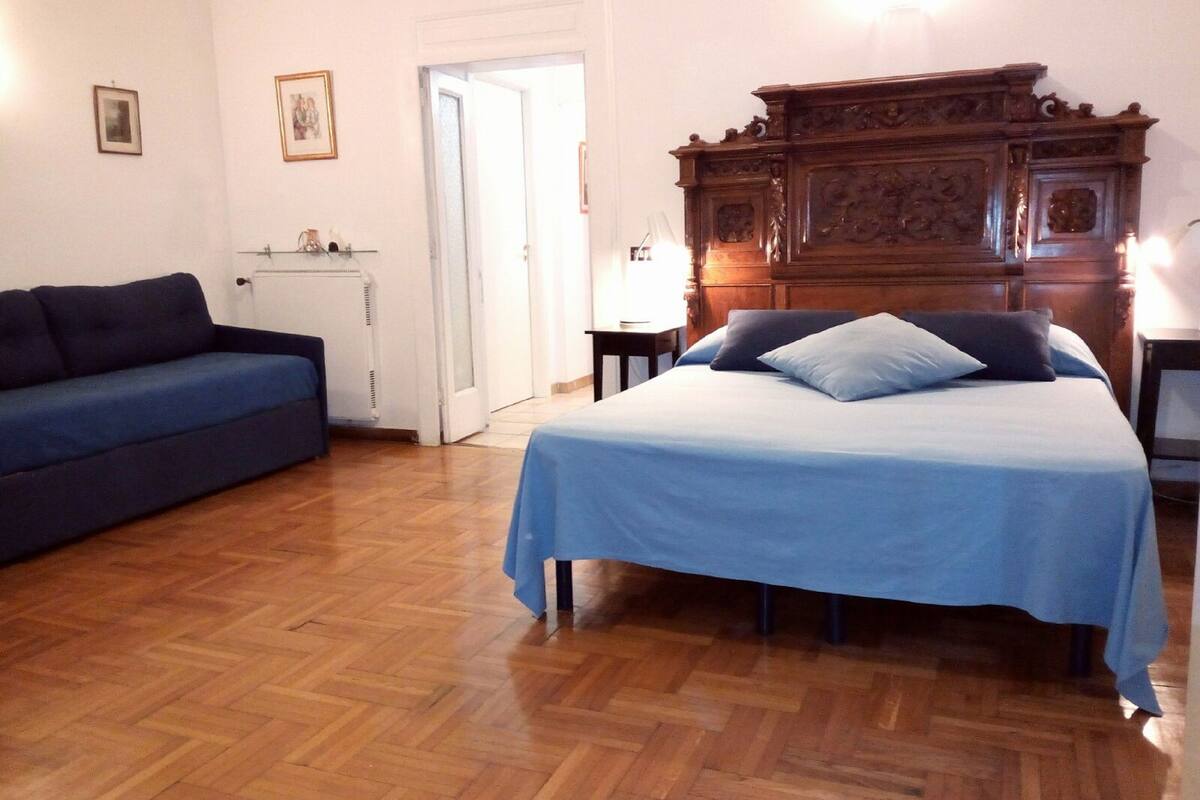 A bedroom with a bed, a wooden dresser, a blue sofa, and a door leading to another room.