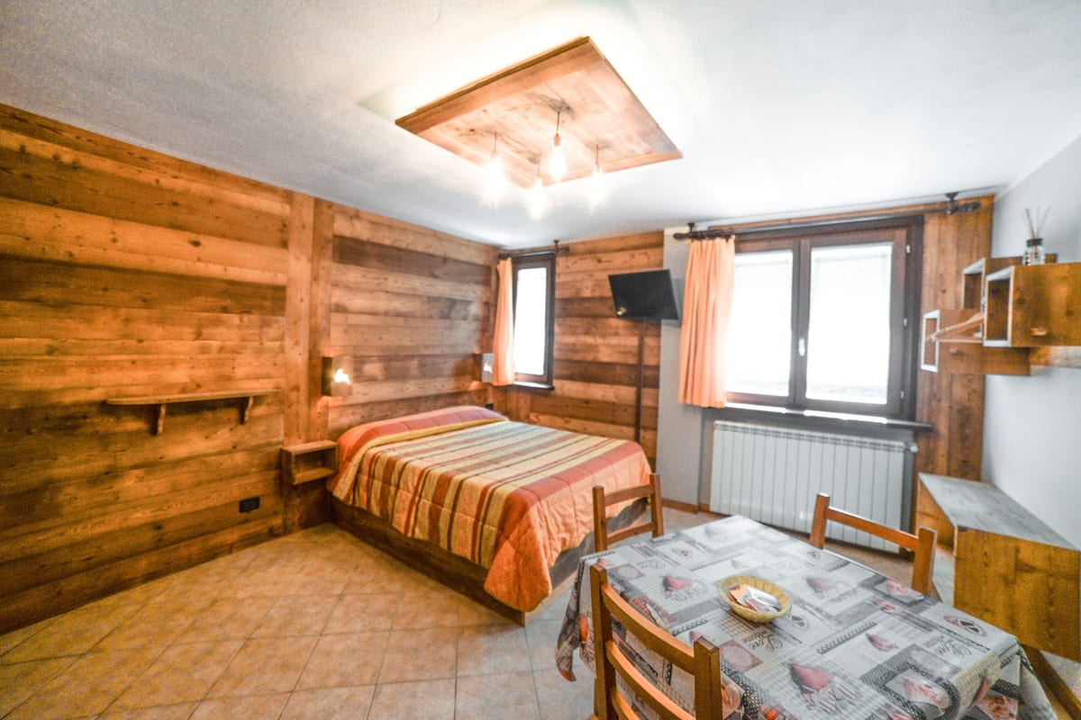 A wooden cabin room with a bed, a table with chairs, a window, and a radiator.