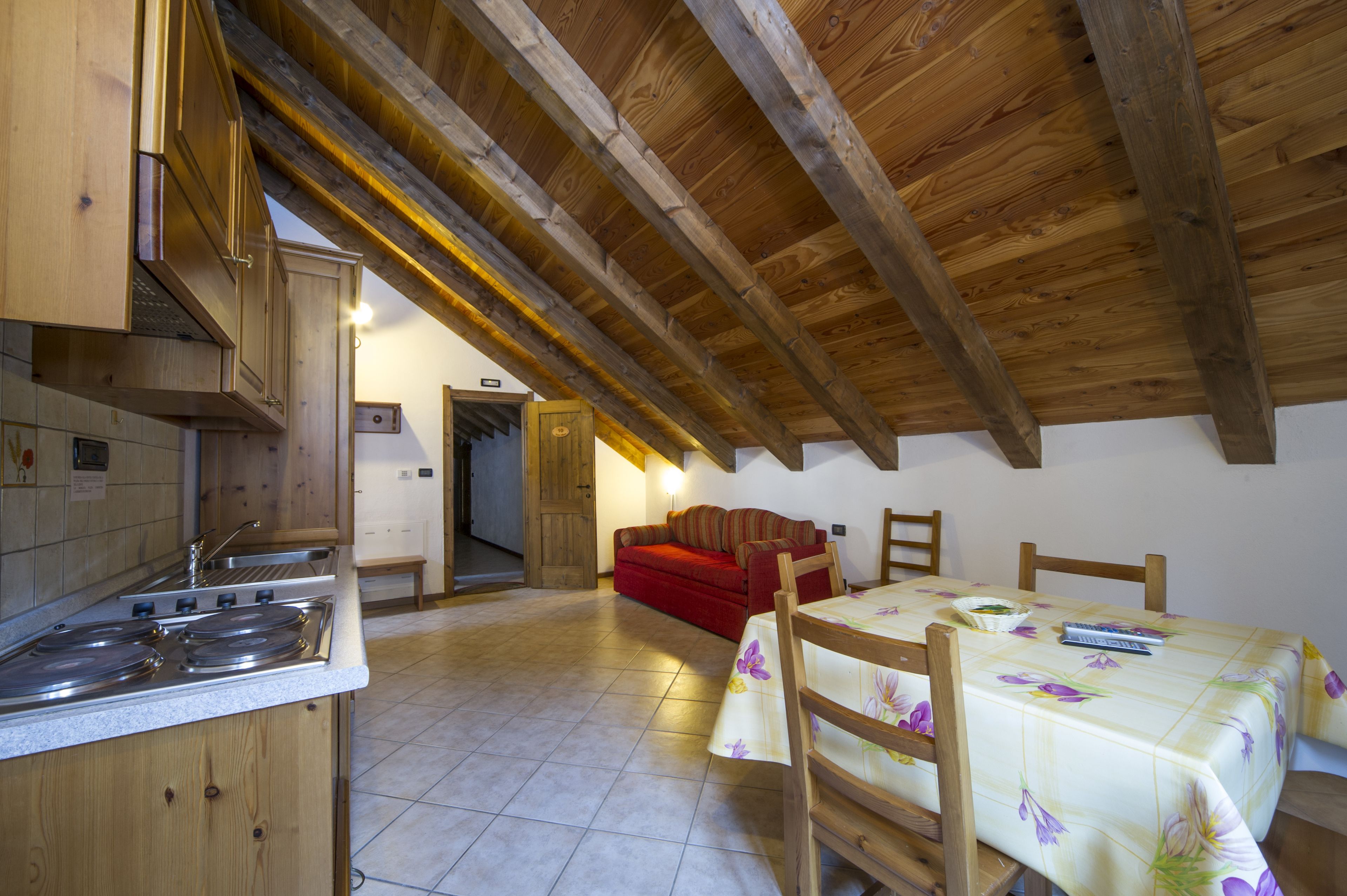 Una cocina con armarios de madera y una estufa, un área de comedor con mesa y sillas, y un sofá rojo en una habitación con techo de madera inclinado.