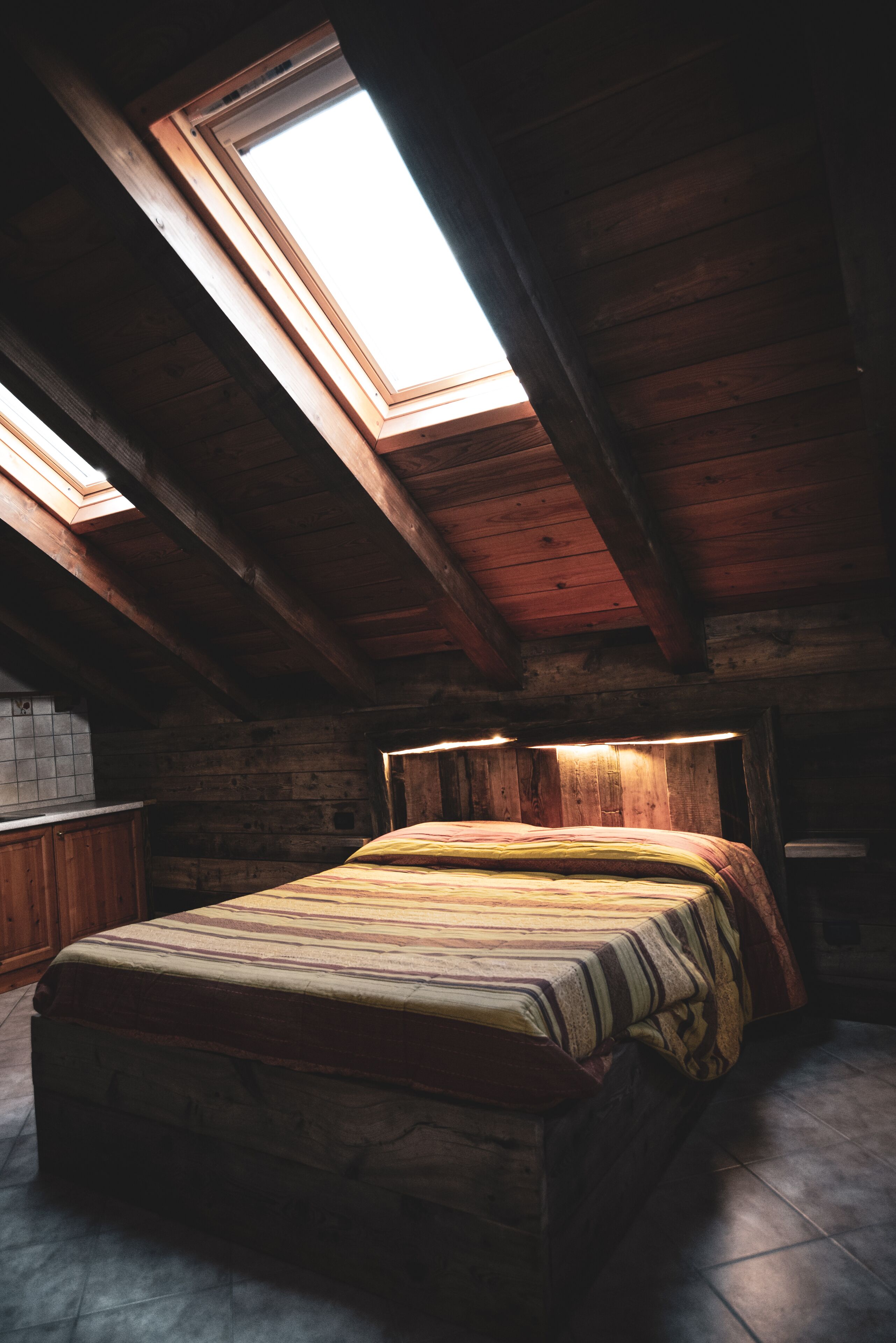 A room with a bed, wooden beams, a skylight, and a tiled floor.