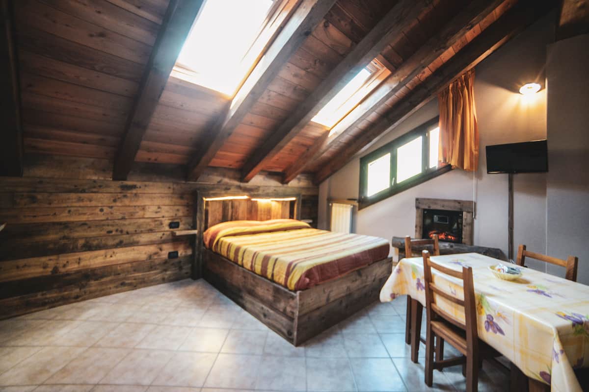 A cozy attic bedroom with a wooden bed, a dining table with chairs, a fireplace, and a skylight.