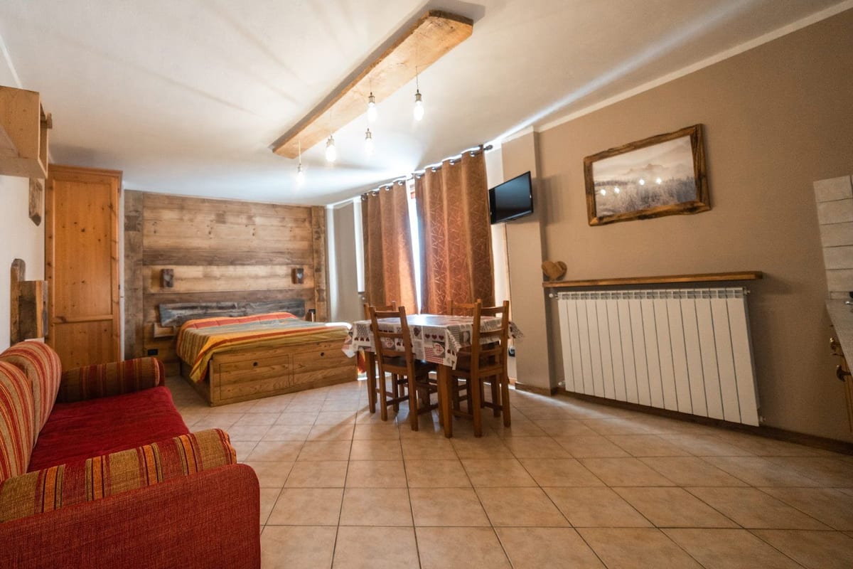 A cozy room with a wooden bed, a sofa, a dining table with chairs, a TV mounted on the wall, and a radiator.