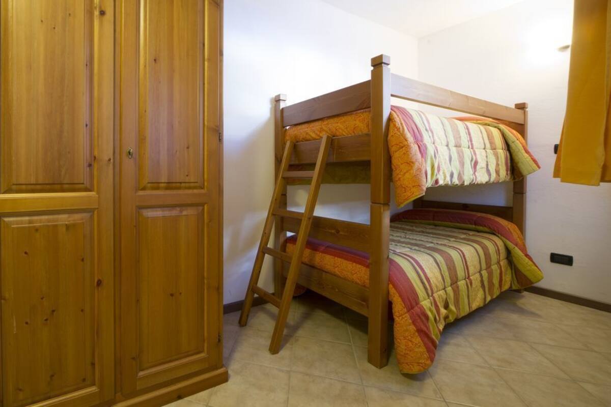 A bunk bed with a wooden ladder, a striped blanket, and a wooden wardrobe in a room with tiled flooring.