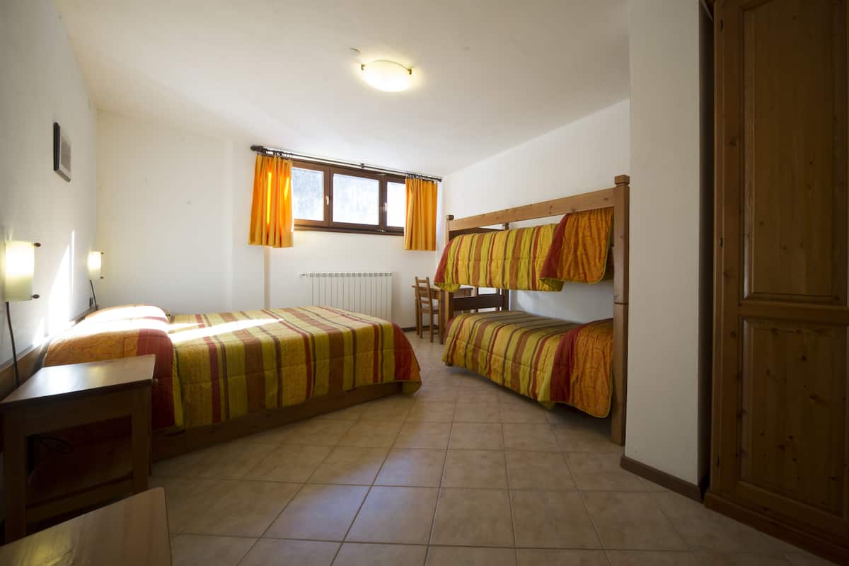 A dormitory room with two beds, a desk, a chair, a radiator, and a window with curtains.