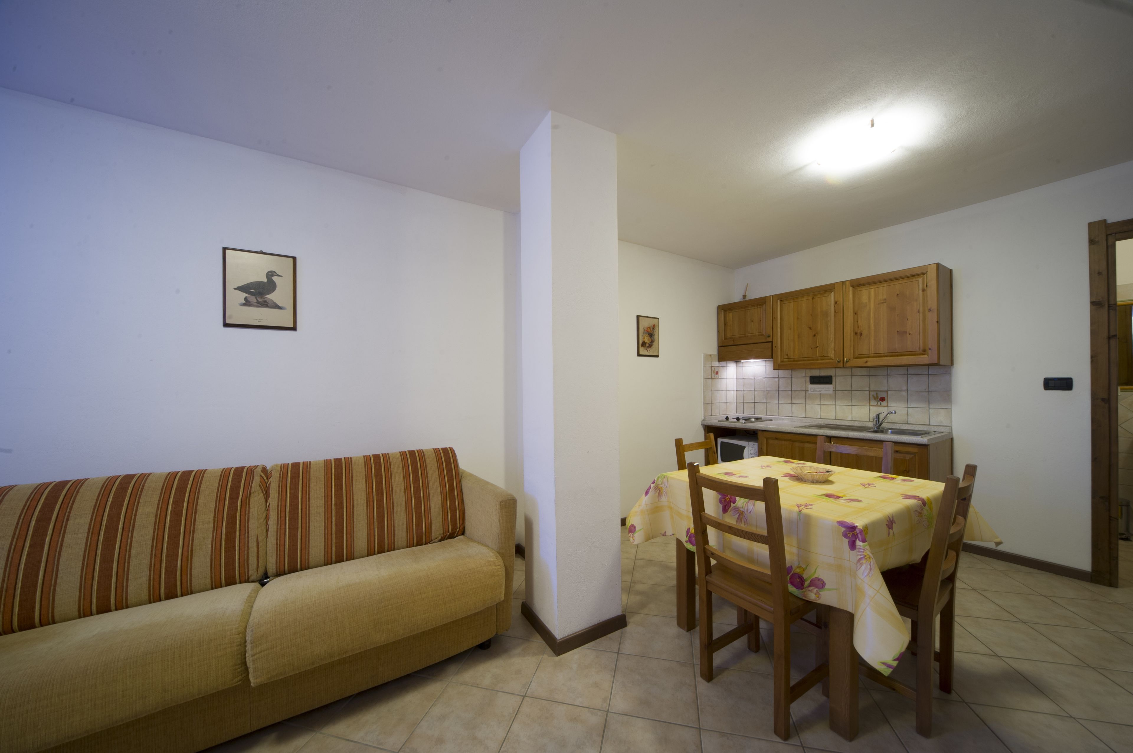 Una sala de estar con sofá y una mesa de comedor con sillas. Se observa una zona de cocina con armarios de madera y salpicadero de azulejos.