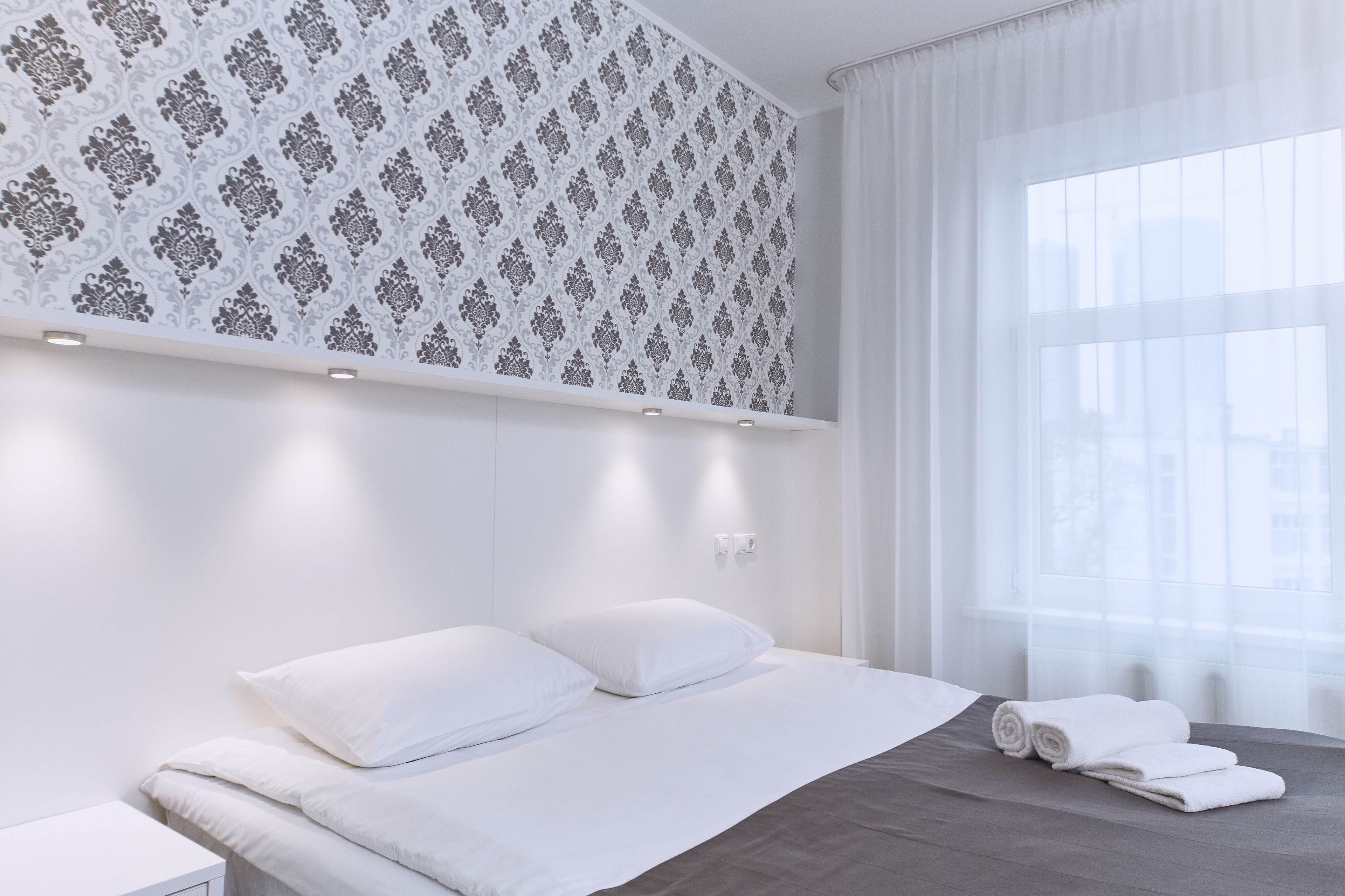A modern hotel room with a large bed, white bedding, a patterned wallpaper, and a large window with sheer curtains.