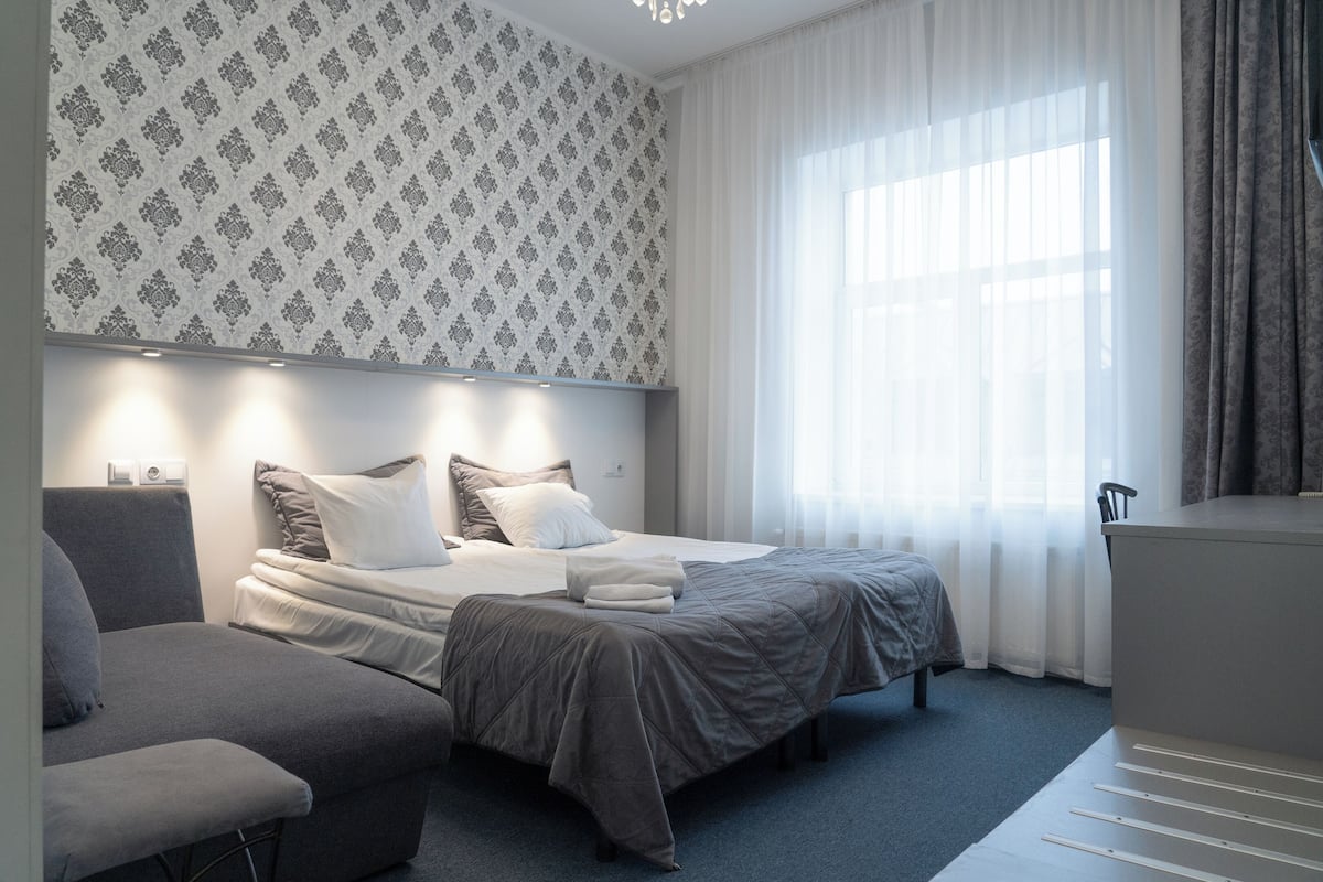 A modern hotel room with a bed, a chair, a desk, and a large window with curtains.
