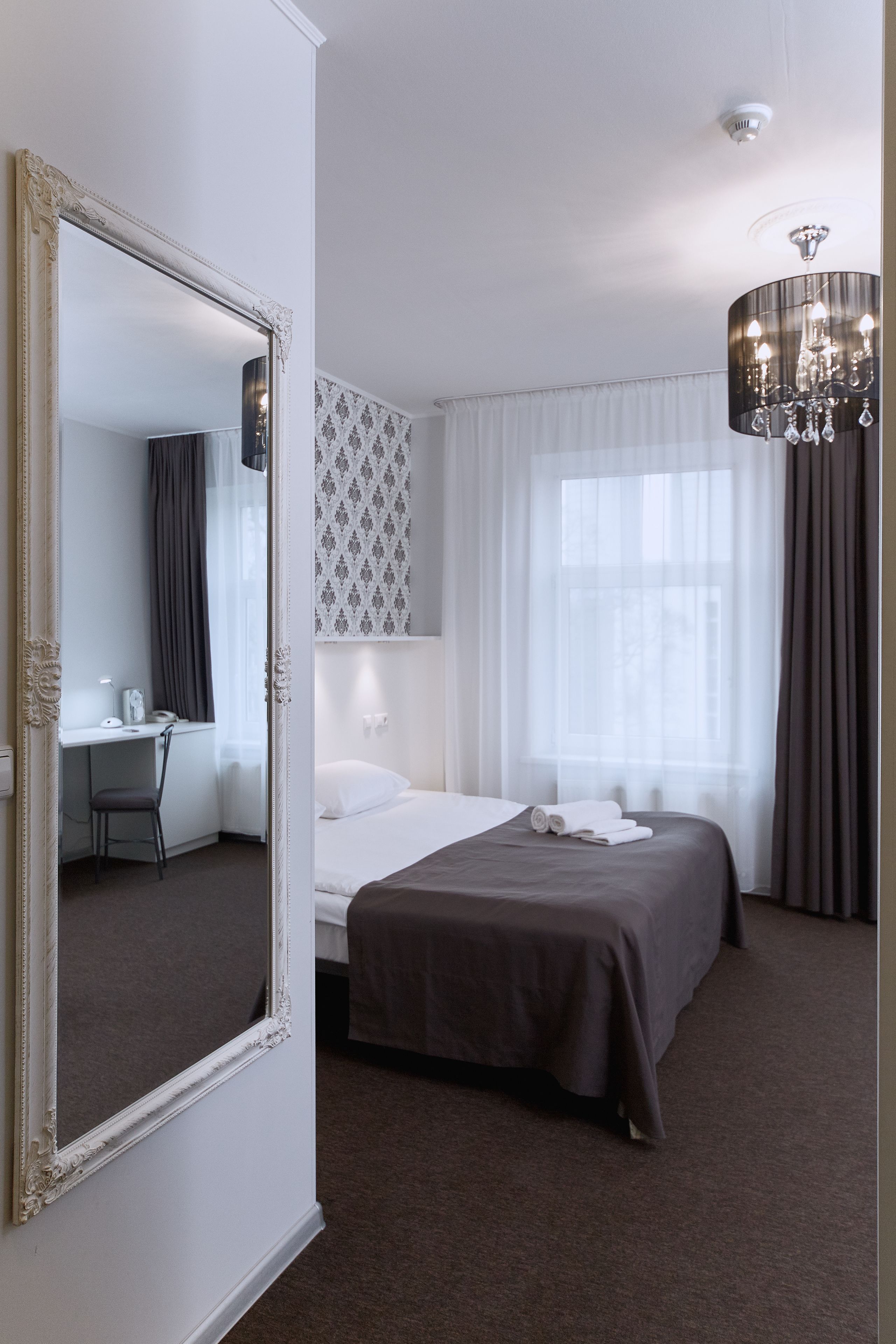 A hotel room with a large mirror, a bed with a dark bedspread, a desk with a chair, and a chandelier.