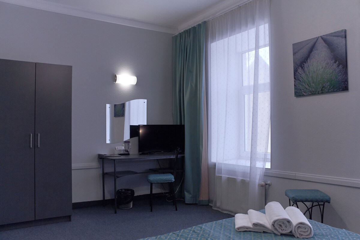 A hotel room with a bed, a desk with a chair, a television, a wardrobe, and a window with curtains.