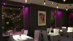 A restaurant interior with tables set for dining, purple ambient lighting, and a view of the street outside. - Hotel OK (Riga)