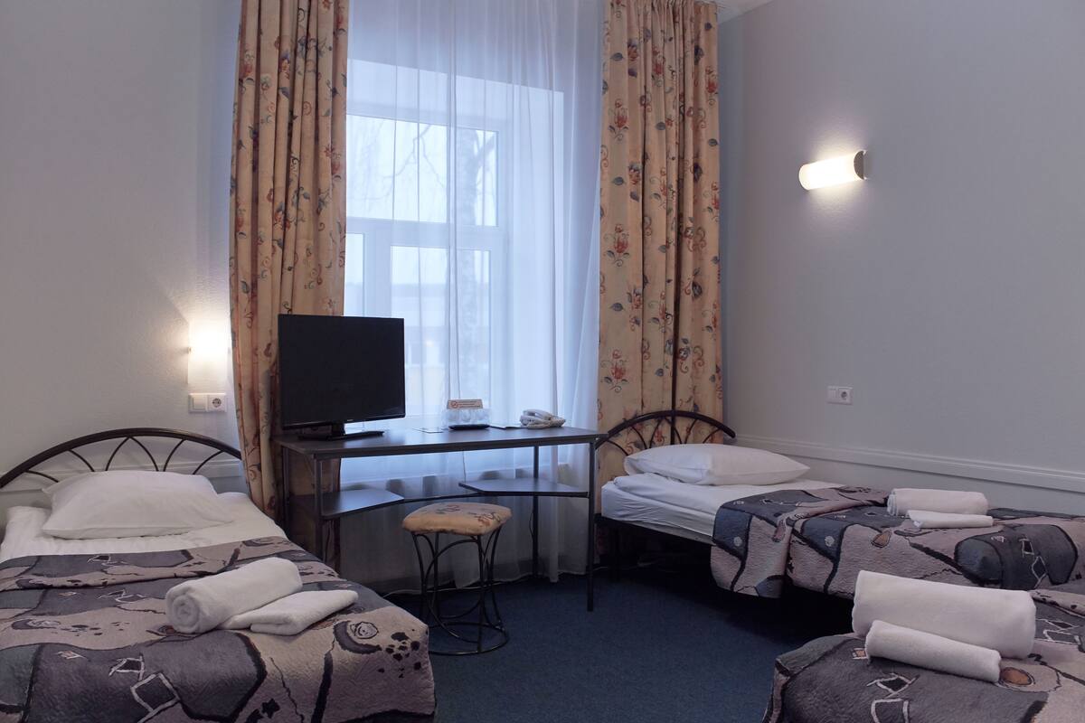 A hotel room with two beds, a desk with a television, and a window with floral curtains.