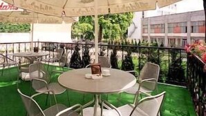 An outdoor patio with round tables and chairs, an umbrella, and a view of buildings and greenery. - Hotel OK (Riga)
