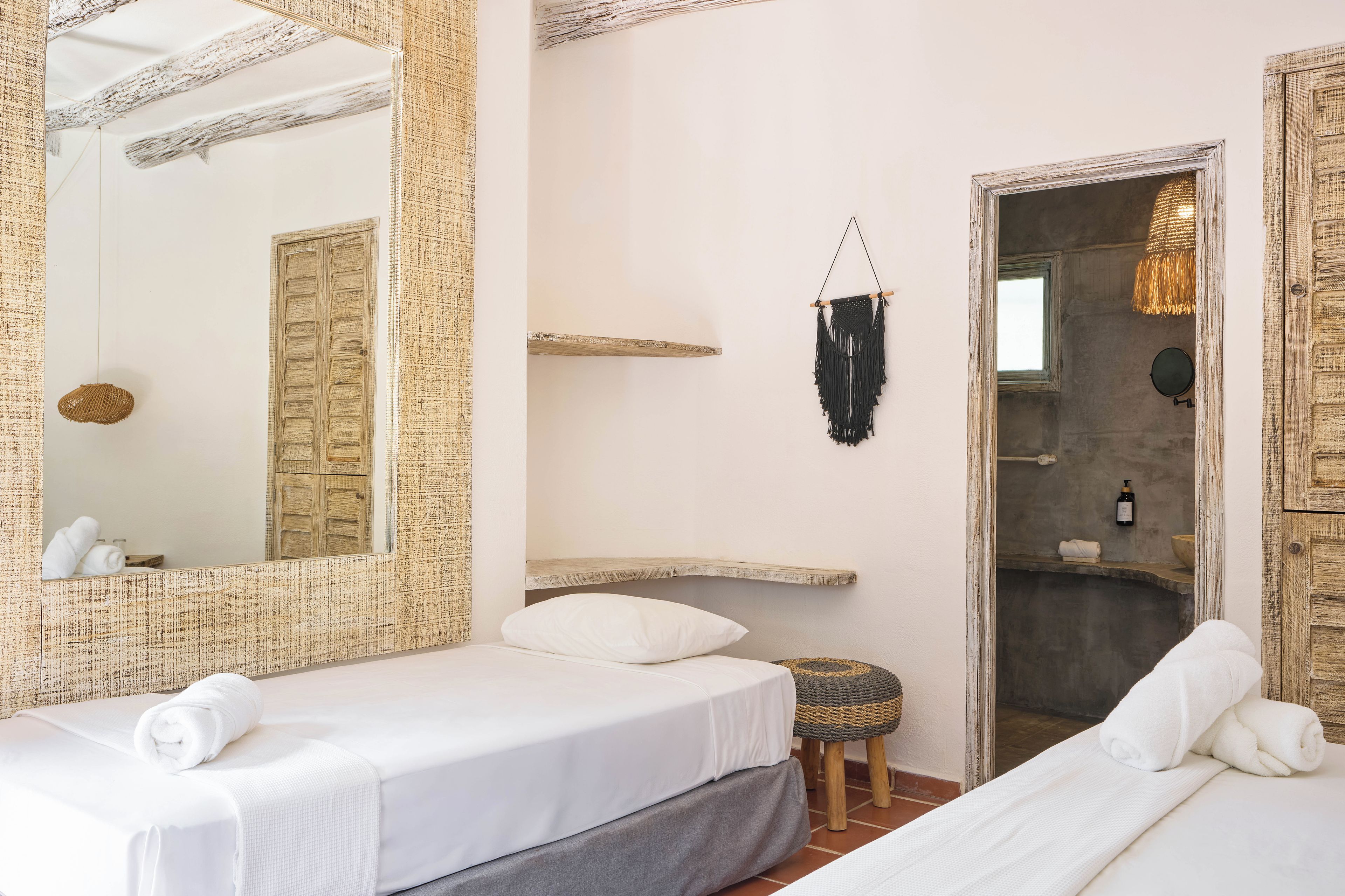 Een hotelkamer met twee bedden, een grote spiegel, een macramé wanddecoratie en een klein krukje.