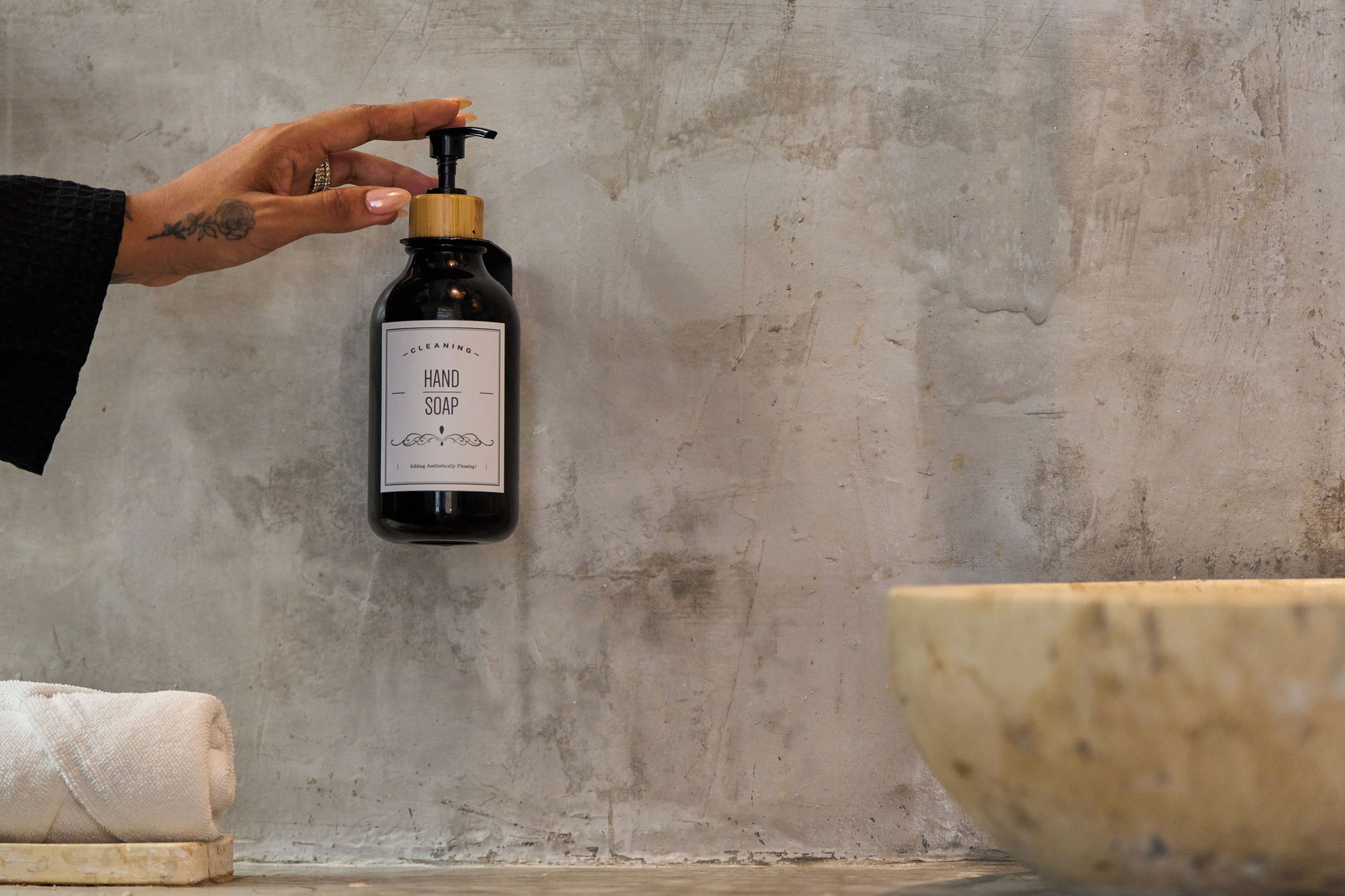 A hand holding a bottle of hand soap with a pump dispenser, a white towel, and a wooden bowl on a concrete surface.