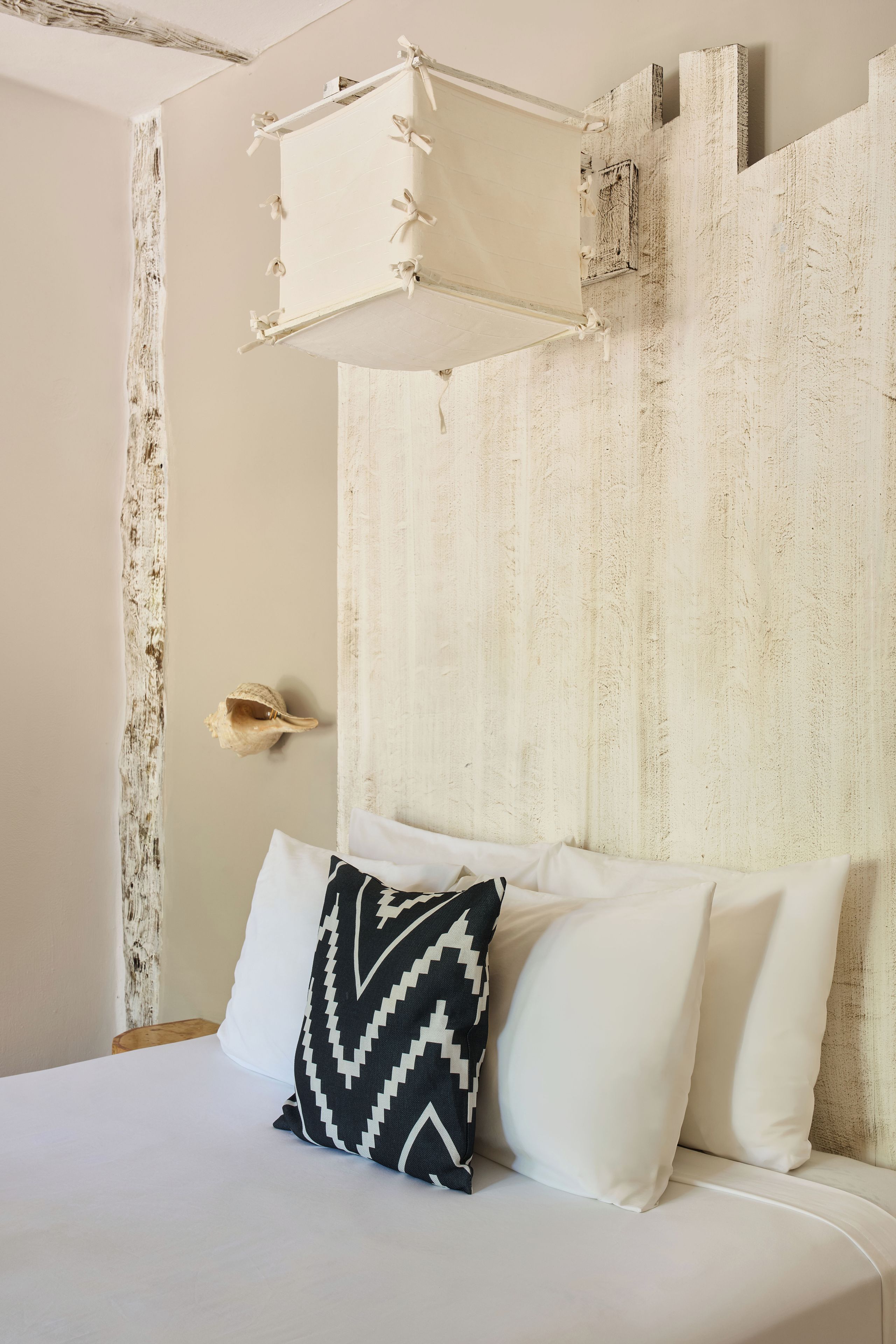 A bed with white bedding and a black and white patterned pillow, a textured wall, and a simple headboard.