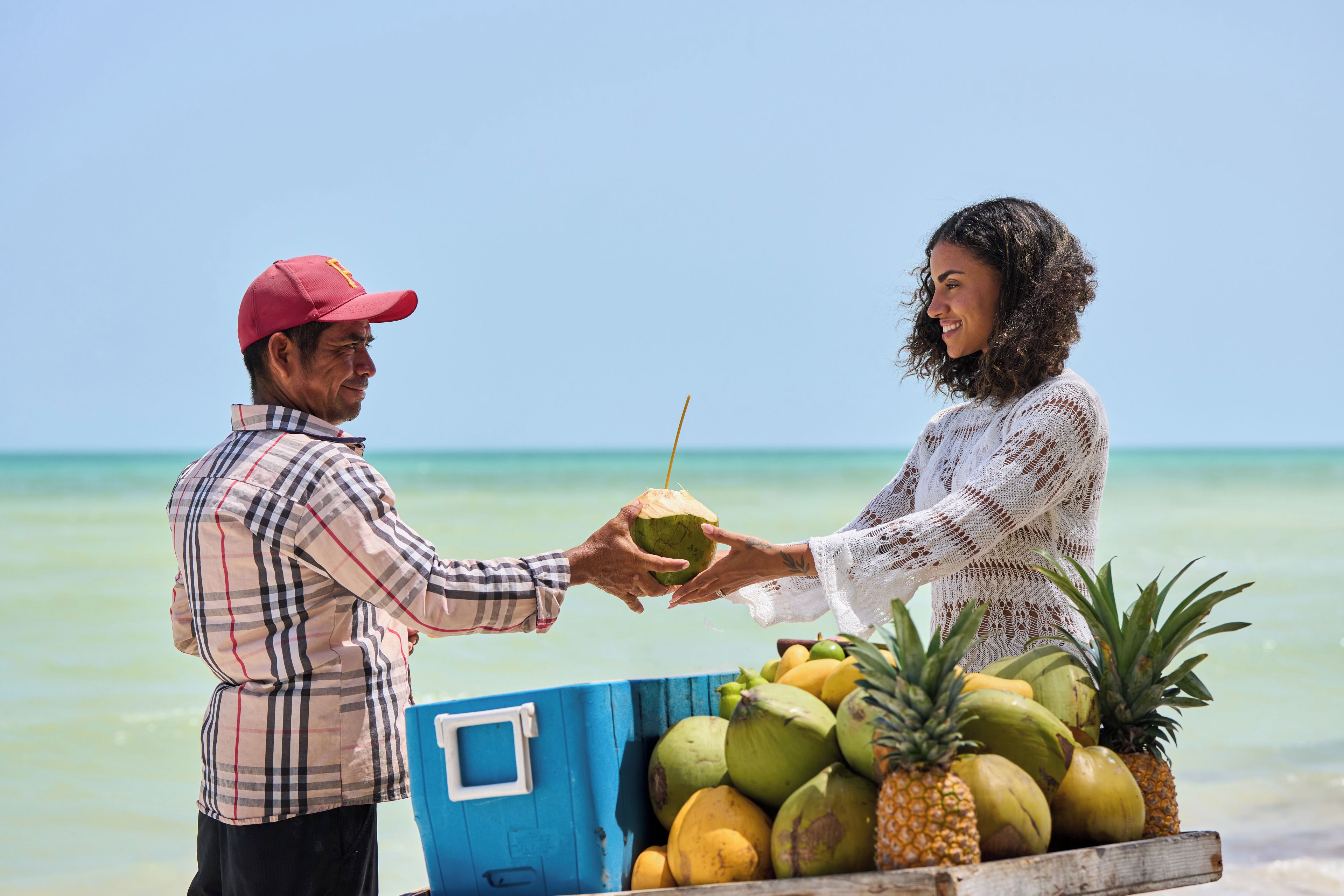 A person handing a coconut to another person on a beach with a blue crate of coconuts and pineapples in the foreground.