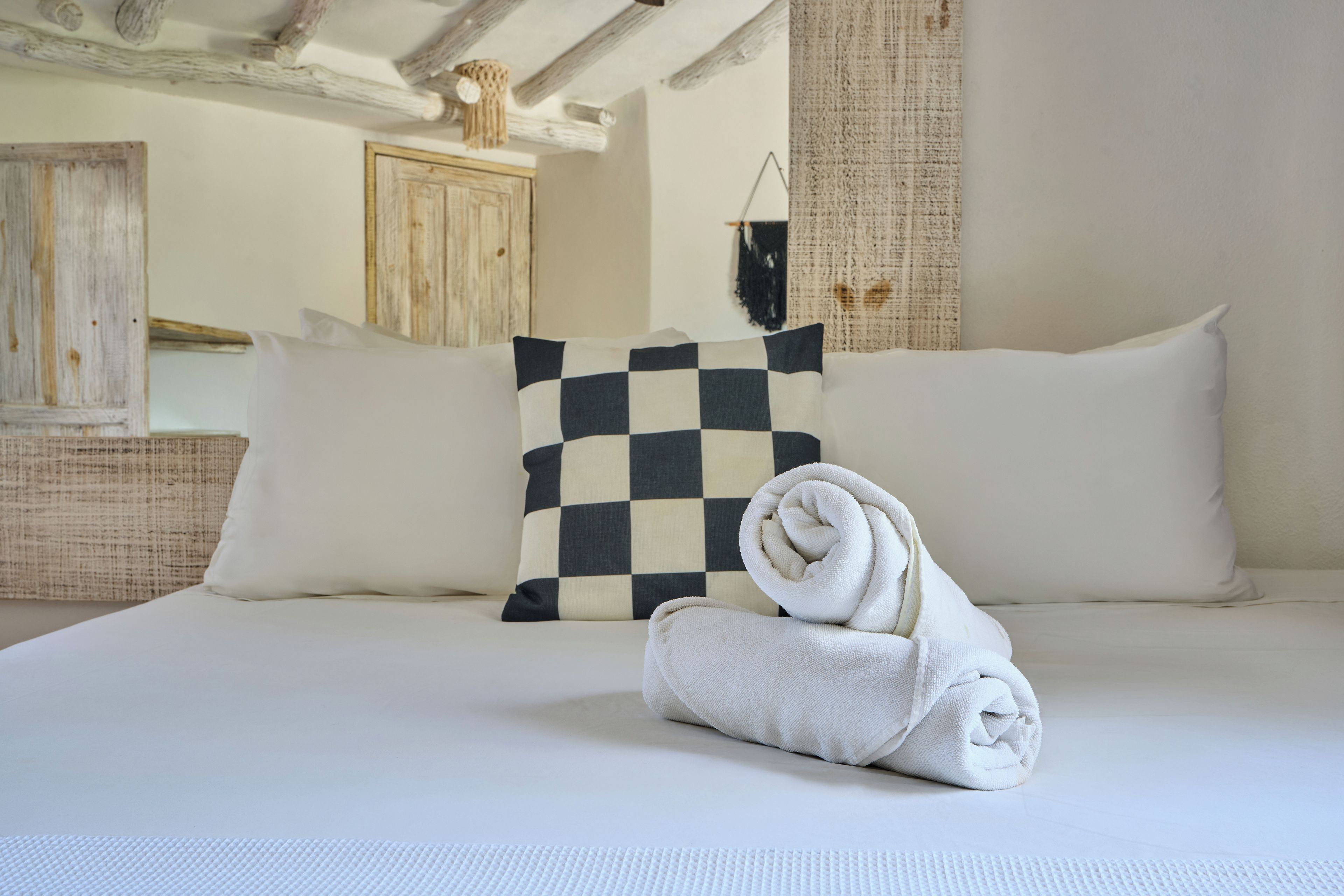 A neatly made bed with a checkered pillow, a rolled white towel, and a textured white mat.