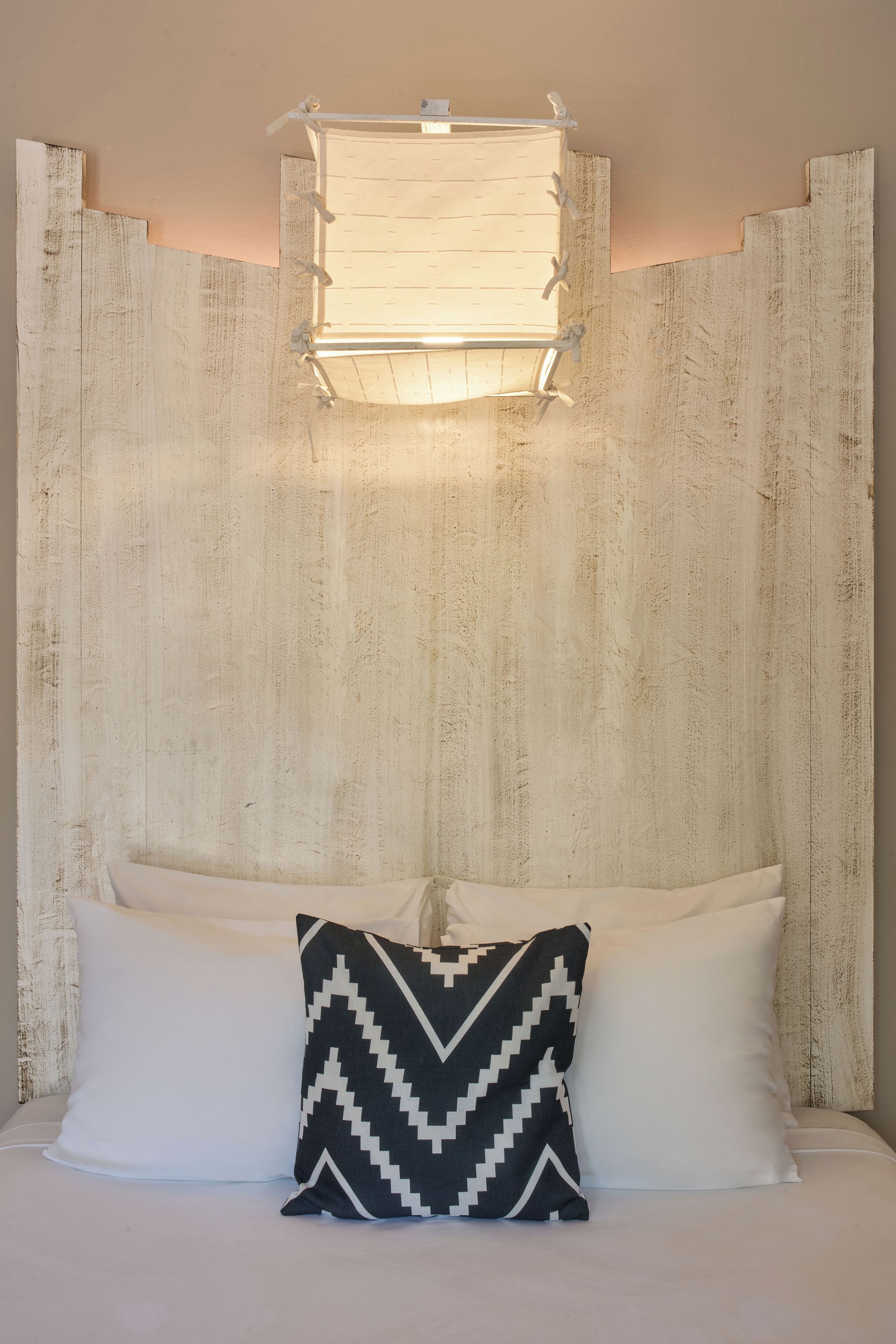 Een bed met twee kussens, waarvan één effen en één met een ruitpatroon, tegen een muren met structuur, met een lamp erboven.