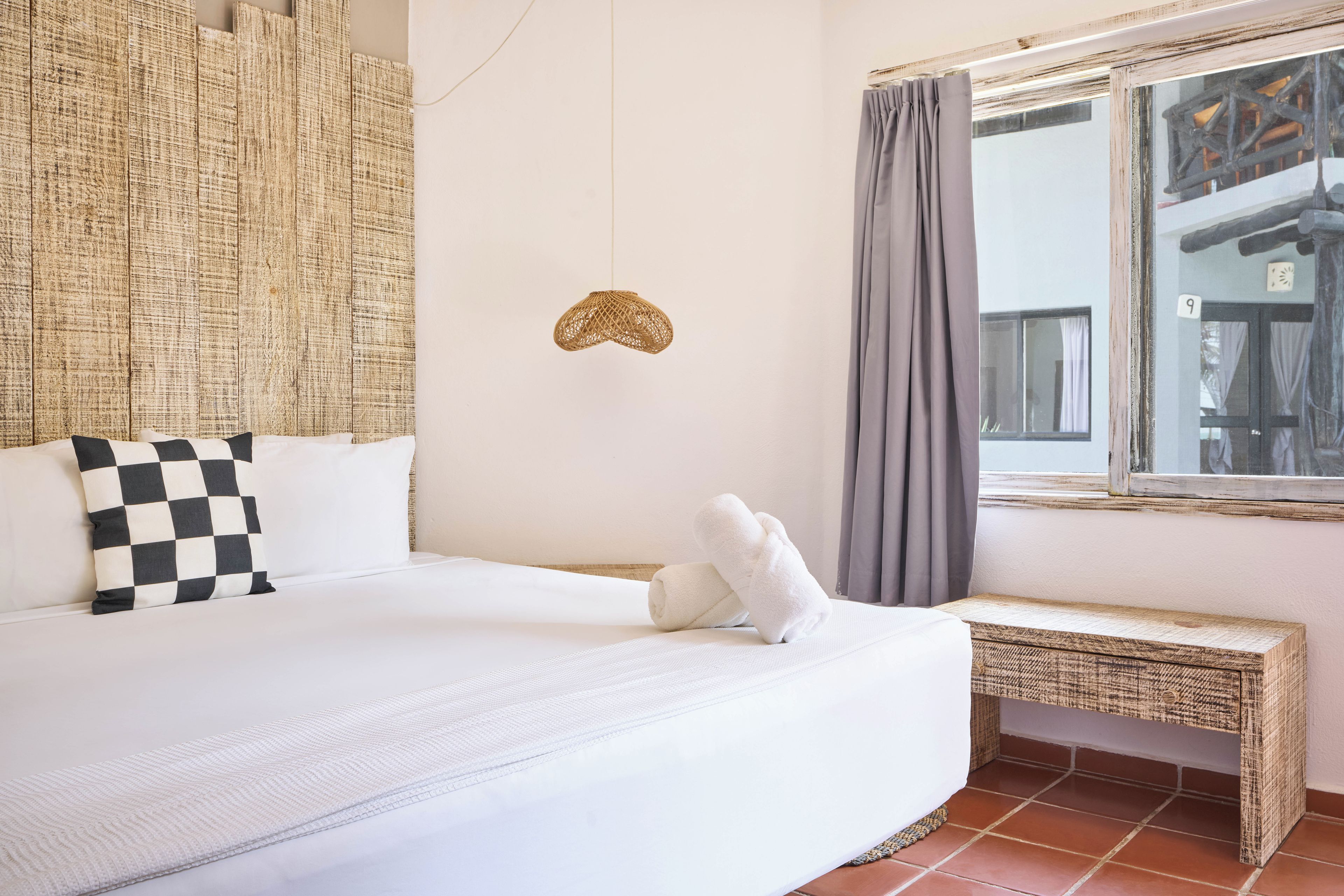 Una habitación de hotel con una cama, almohada de cuadros, cabecero tejido, una pequeña decoración en la pared, una ventana con cortinas y un banco.