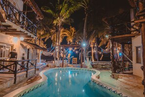 Un área de piscina nocturna con temática tropical, que incluye palmeras y una estructura con techo de paja.