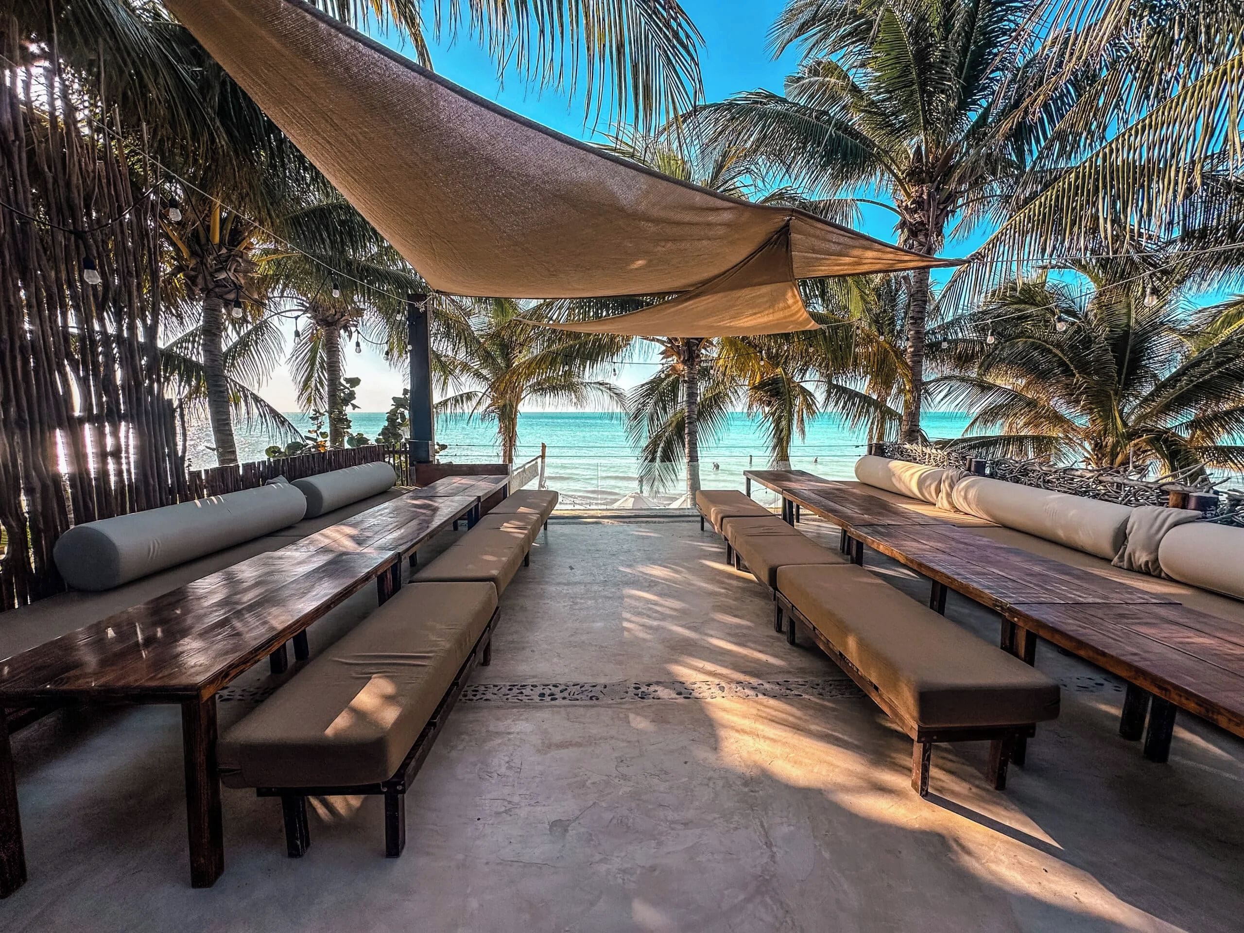 Un área de estar frente a la playa con mesas y bancos de madera, un toldo arriba y vista al océano.