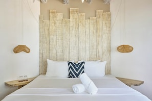 Une chambre d’hôtel avec un grand lit, une tête de lit à motifs, deux lampes fixées au mur et une petite table d’appoint en bois sur laquelle sont posés des verres.