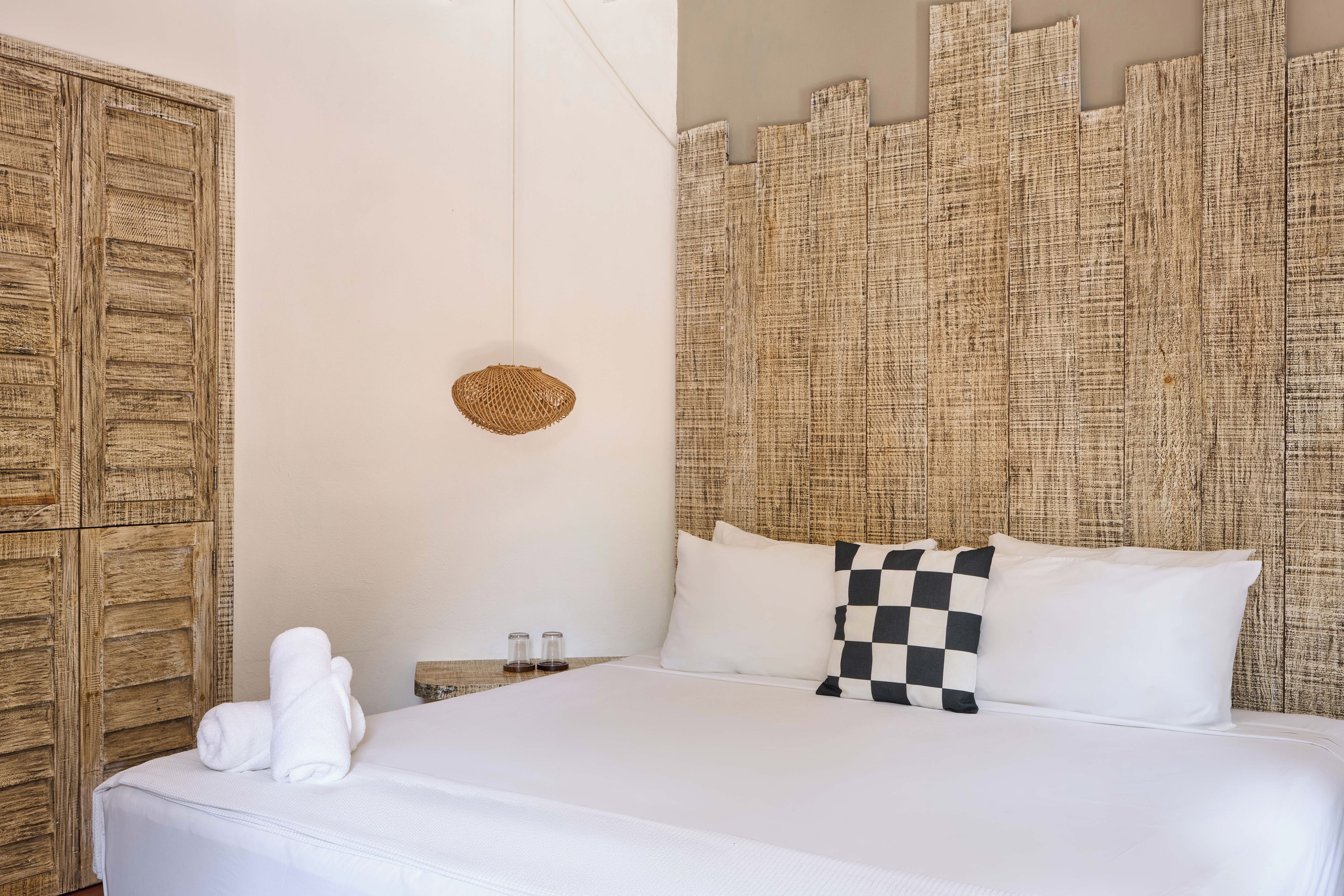Habitación de hotel con una cama grande, cabecera de madera, mesita de noche pequeña con salero y pimentero, y una toalla blanca sobre la cama.