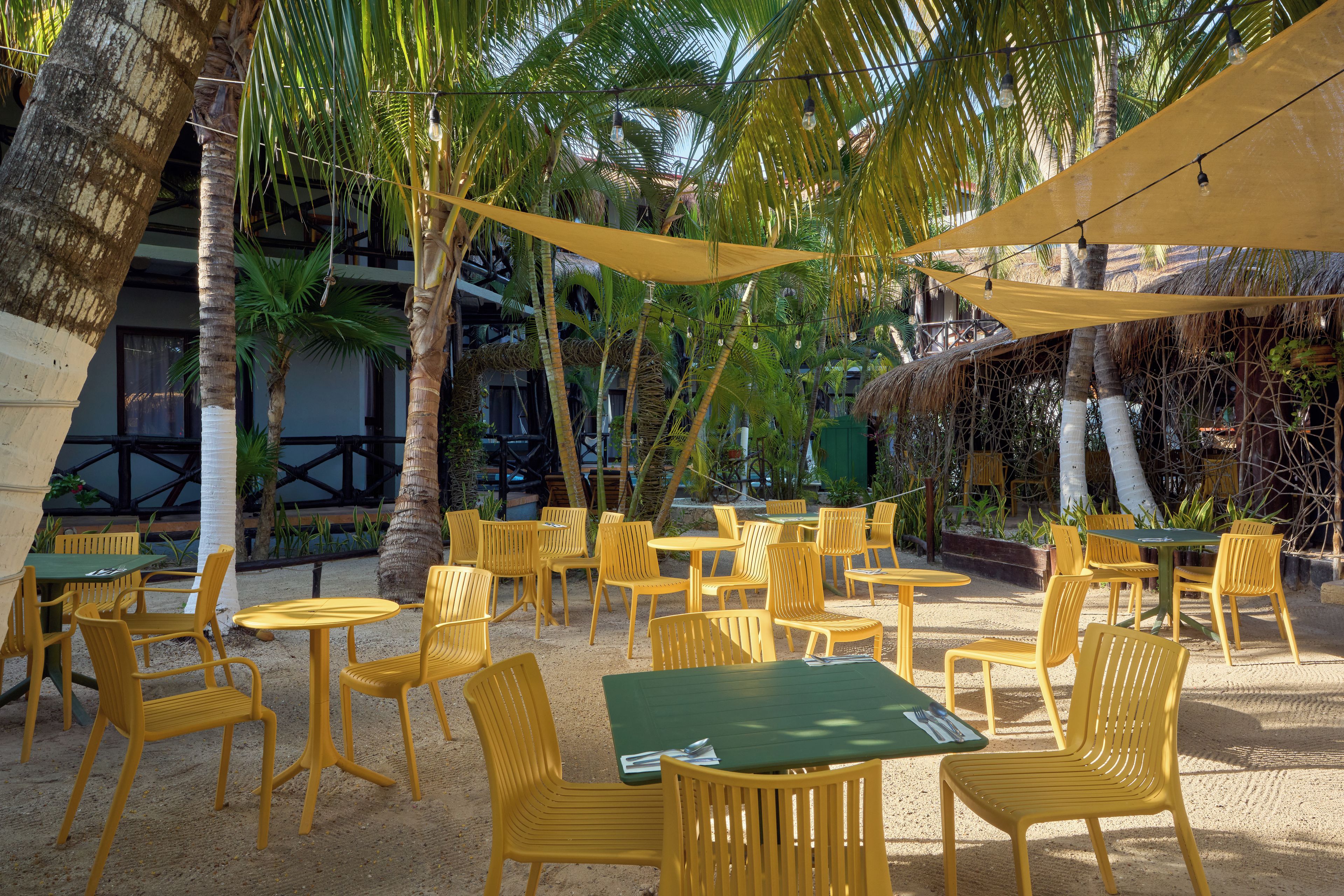 Een terras met gele stoelen en groene tafels, omgeven door palmbomen en een constructie met rieten dak.