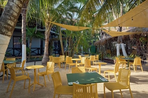 An outdoor seating area with yellow chairs and green tables, surrounded by palm trees and a thatched-roof structure.