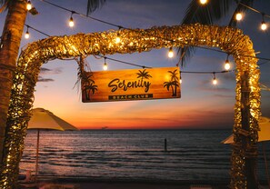 Señal de un club de playa con palmeras, rodeado de luces colgantes y vistas al atardecer.