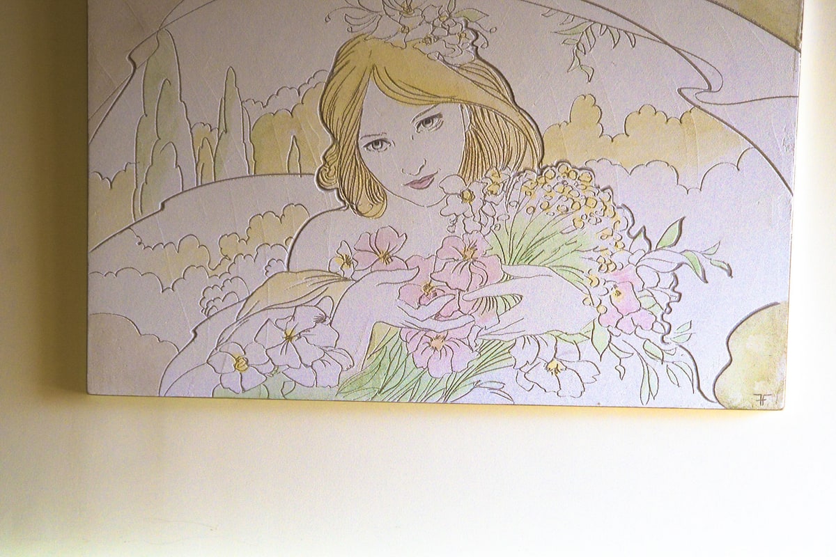 A wall-mounted artwork of a woman with flowers, a bed with a patterned headboard, and a plain wall.