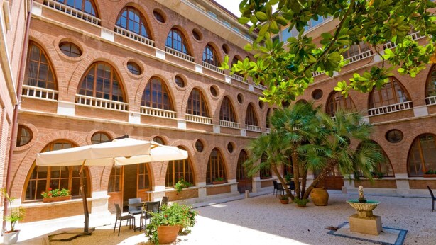 A courtyard with a brick building featuring arched windows and balconies, outdoor seating, and potted plants. - San Francesco Hotel (Loreto)