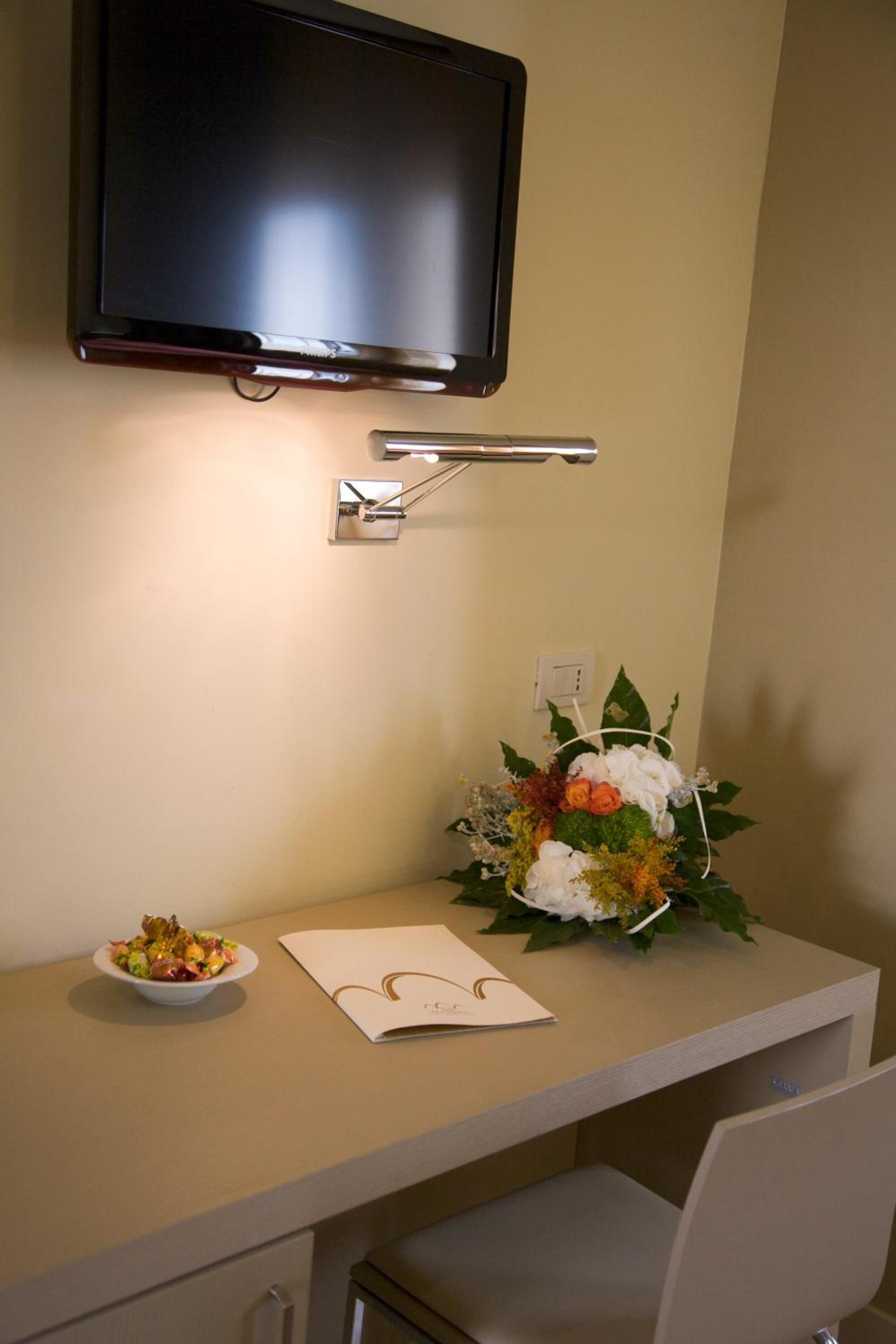 A hotel room with a flat-screen TV mounted above a desk, a small table with a bowl of fruit, a bouquet of flowers, and a piece of paper.