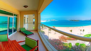 A balcony with a view of a beach, green lounge chairs, and a wooden table.