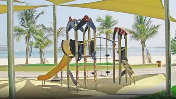 Explore the playground's climbing and sliding elements under a shaded canopy amidst palm trees and a beach.