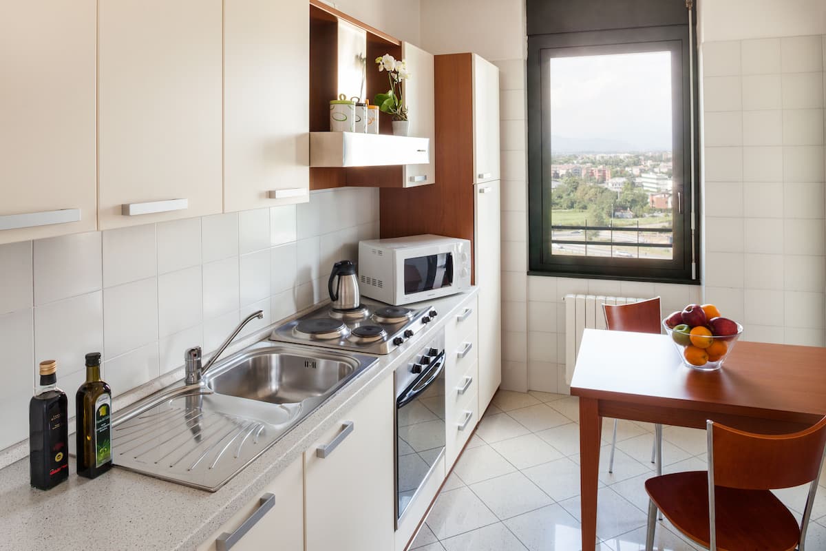 Une cuisine moderne avec des armoires blanches, un évier en acier inoxydable, un four à micro-ondes et une cuisinière. Une fenêtre offre une vue sur les bâtiments, et un saladier rempli de fruits est posé sur la table.